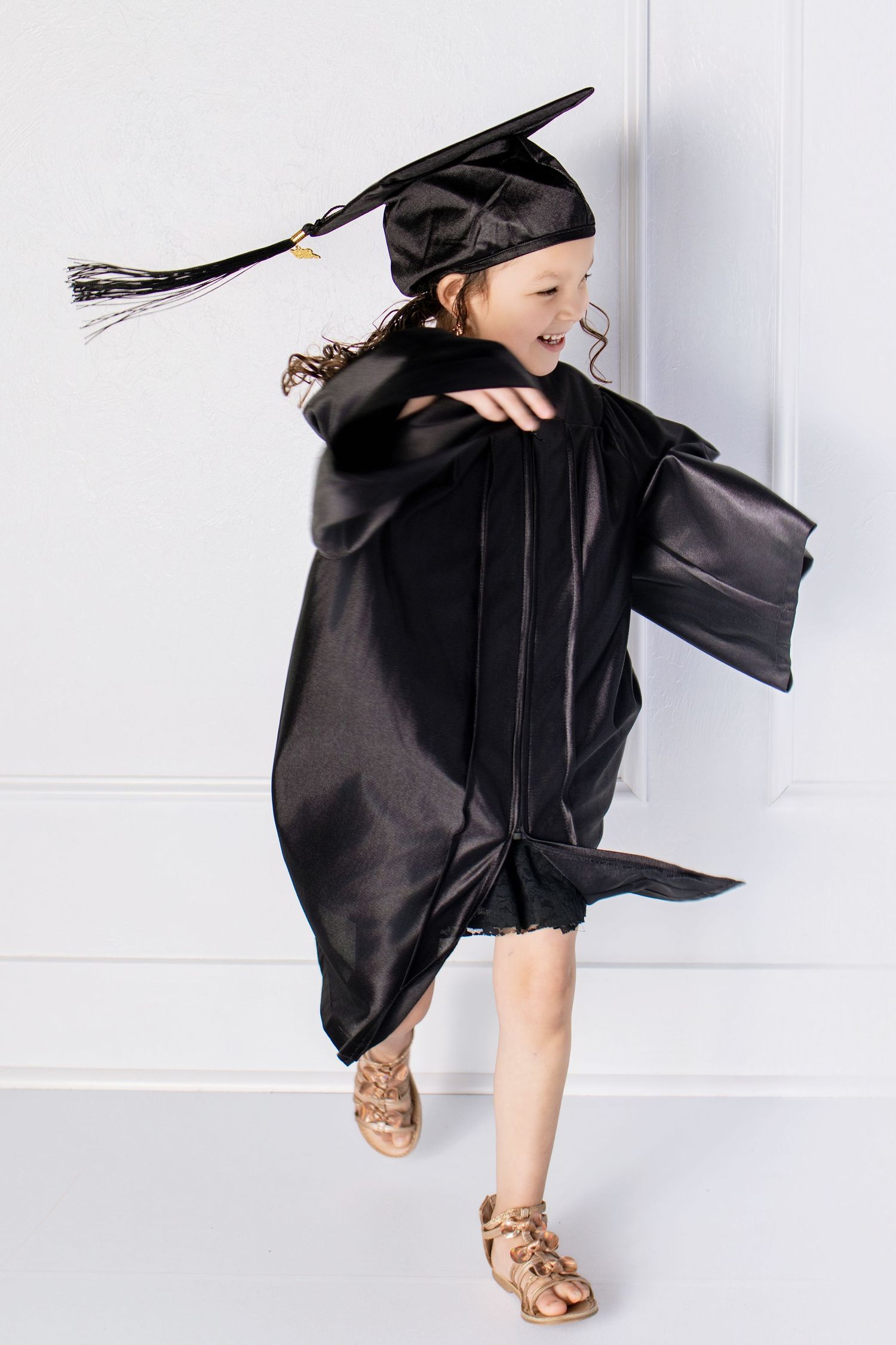 Joyful little girl in black graduation cap and gown spinning and twirling against a white background.