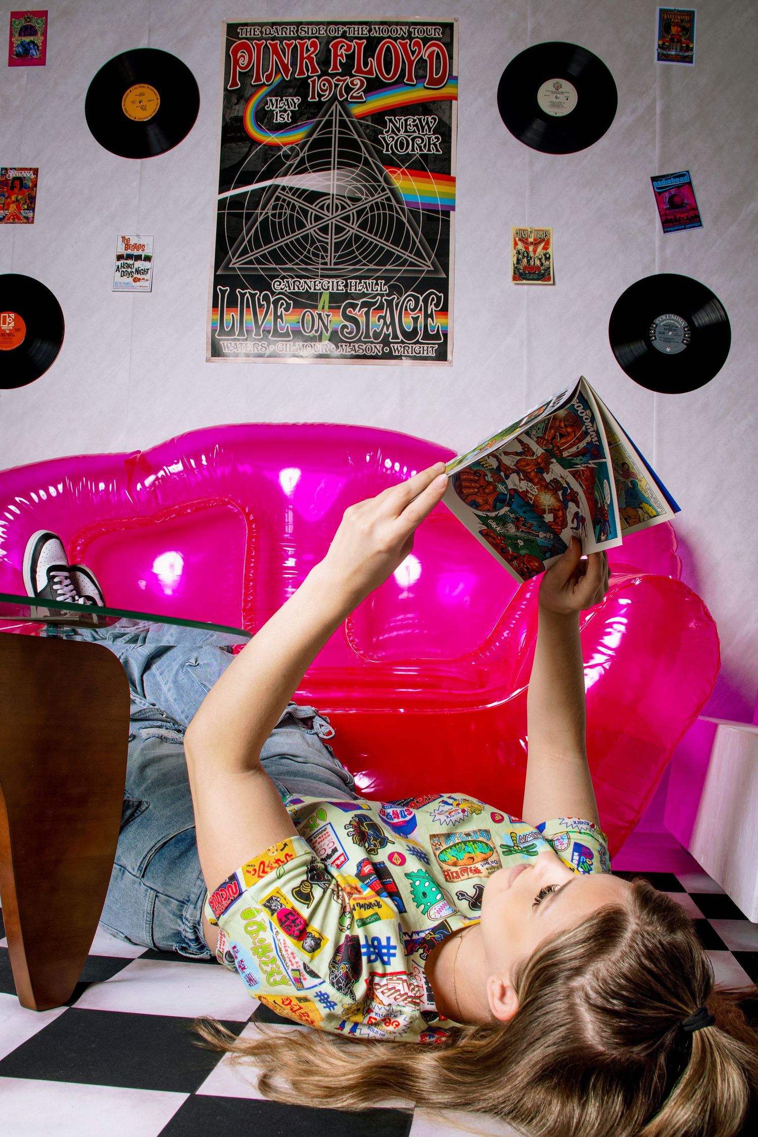 Teen girl in graphic shirt reads comic book while lying on pink inflatable sofa near Pink Floyd poster.