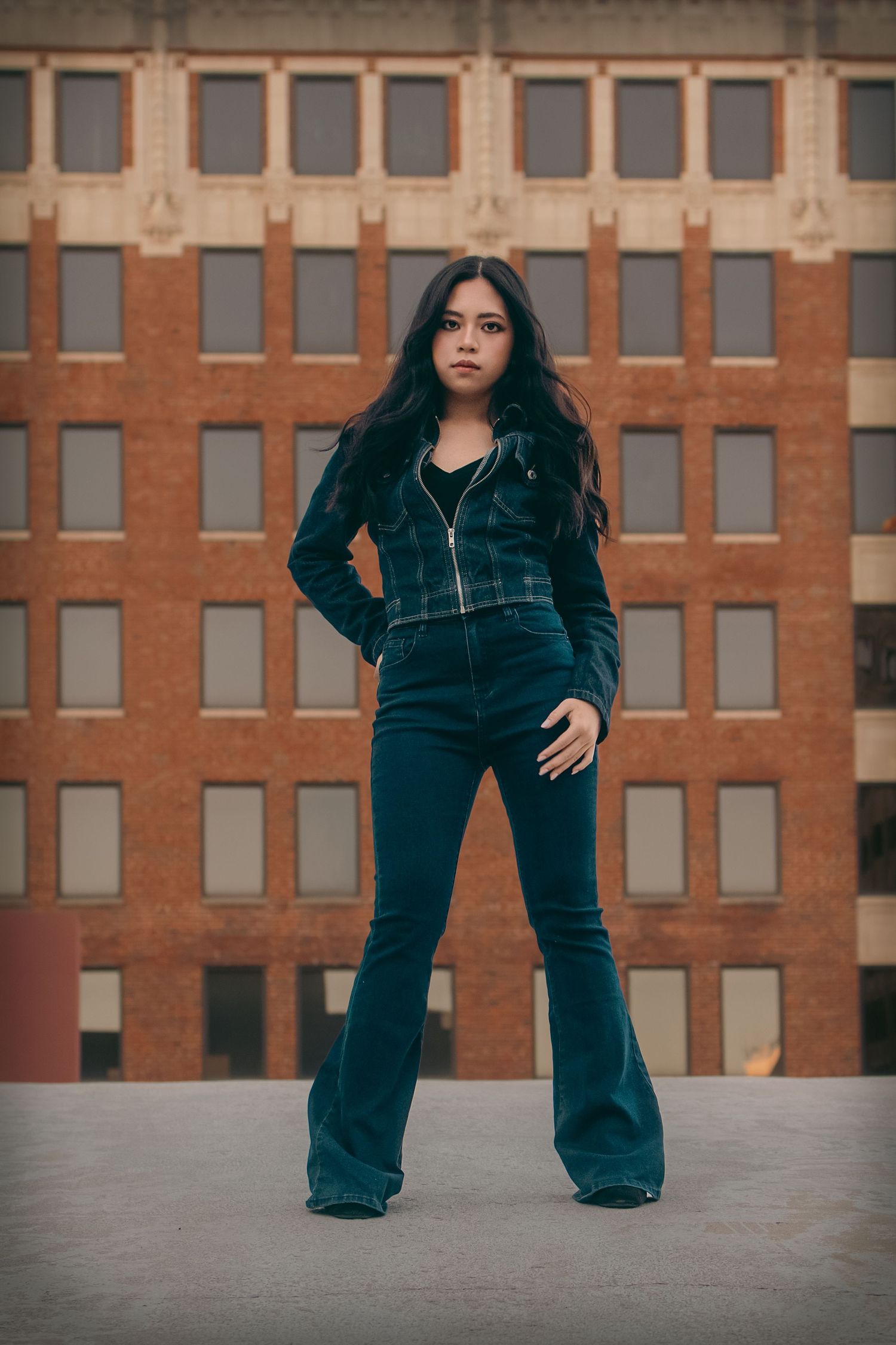 Woman in teal plaid jacket and flare jeans poses confidently in front of brick building.