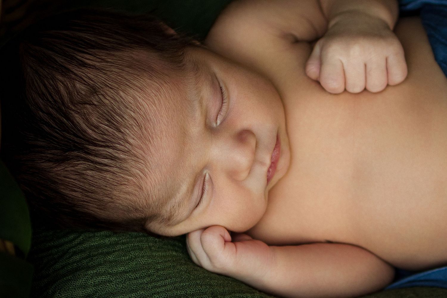 Peaceful newborn baby sleeping curled up on a green blanket, with tiny fingers and dark hair visible.