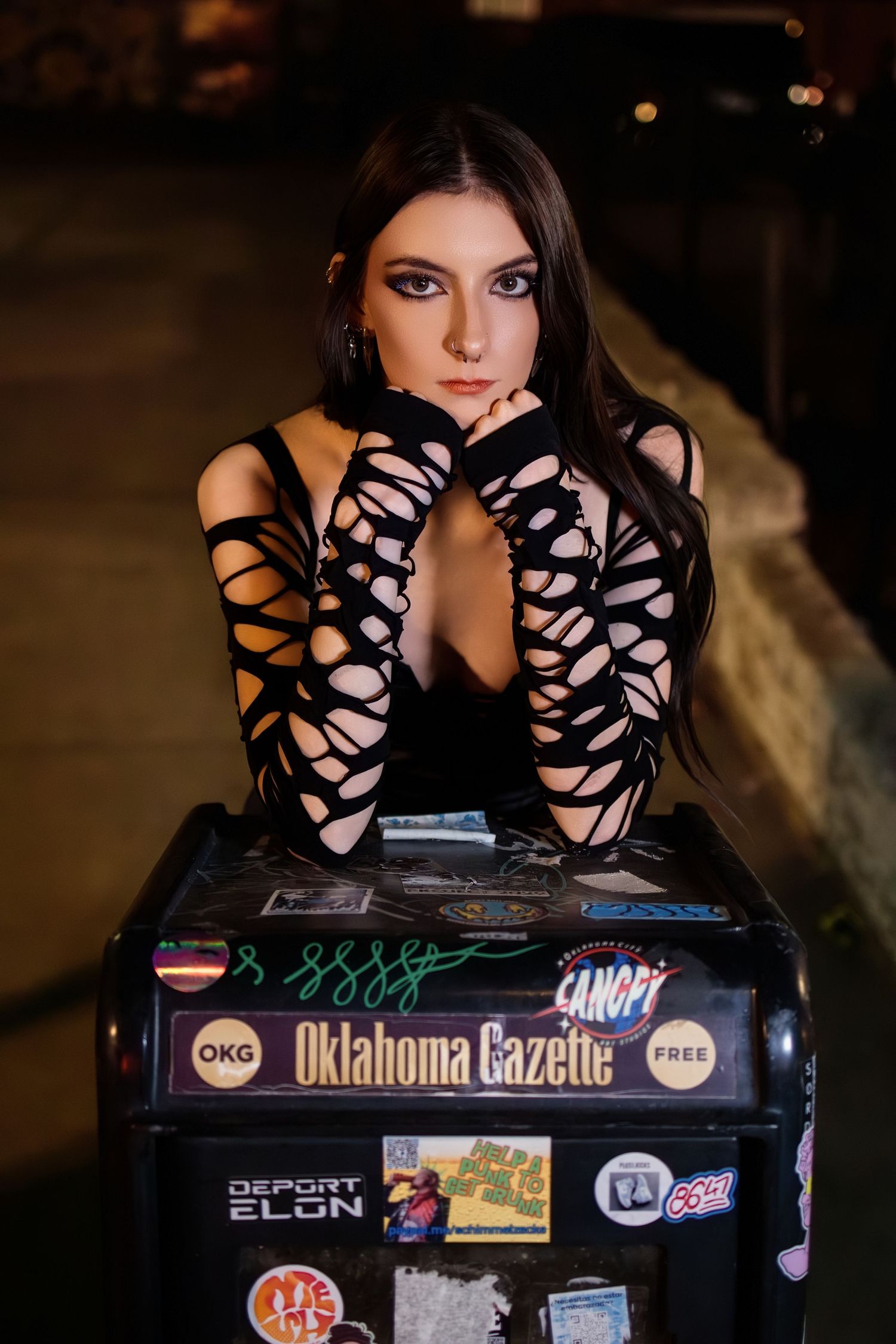 Young woman in black cutout gloves poses on Oklahoma Gazette newspaper box with stickers at night.