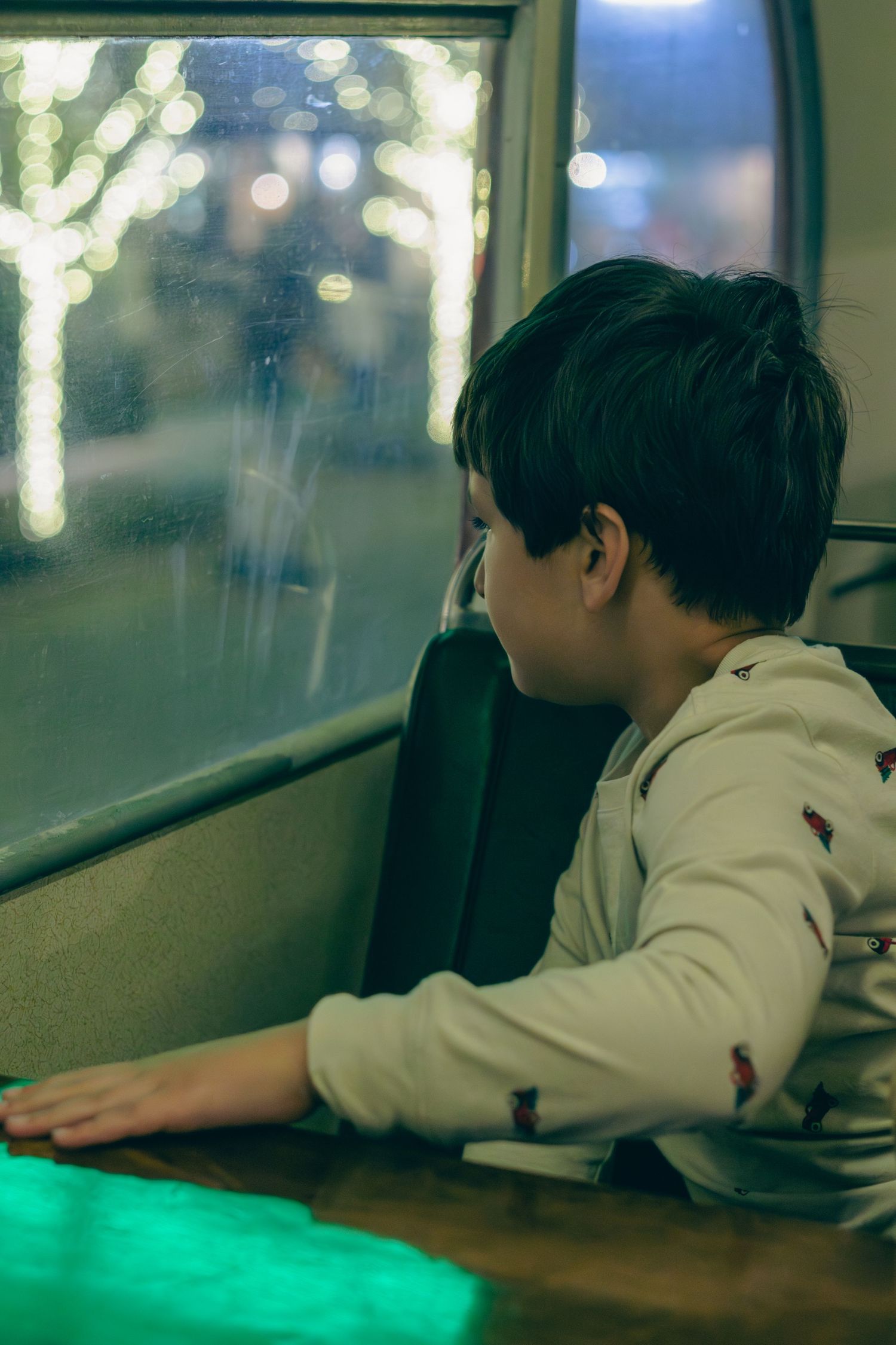 A young boy gazes thoughtfully out a rain-streaked window at night, seated in a vehicle with bokeh lights outside.