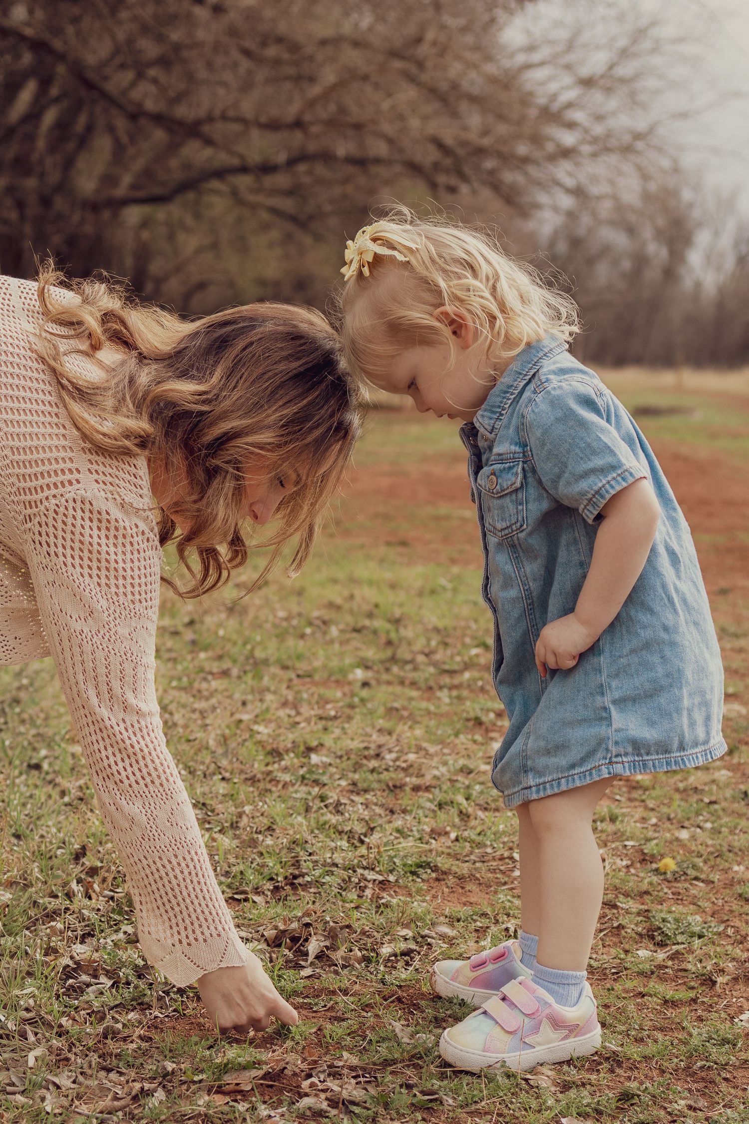 Mother and toddler girl in denim dress touching heads together outdoors in a park setting.