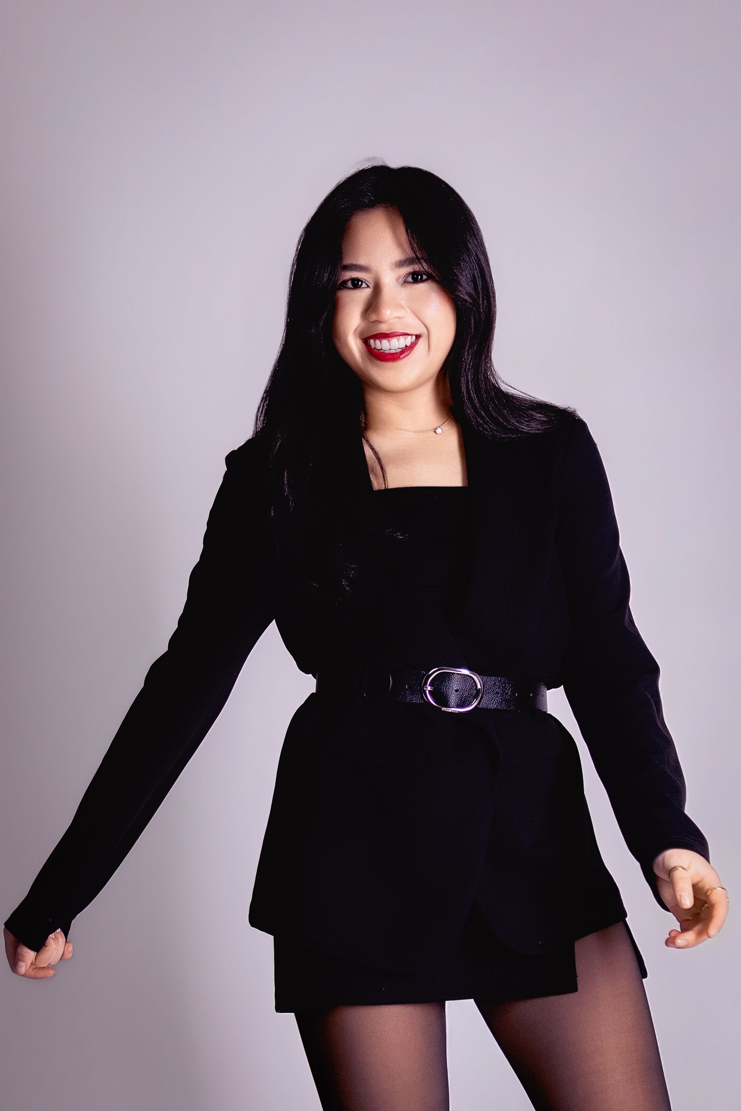 Young woman in black belted blazer dress with red lipstick, smiling and posing against a gray background.