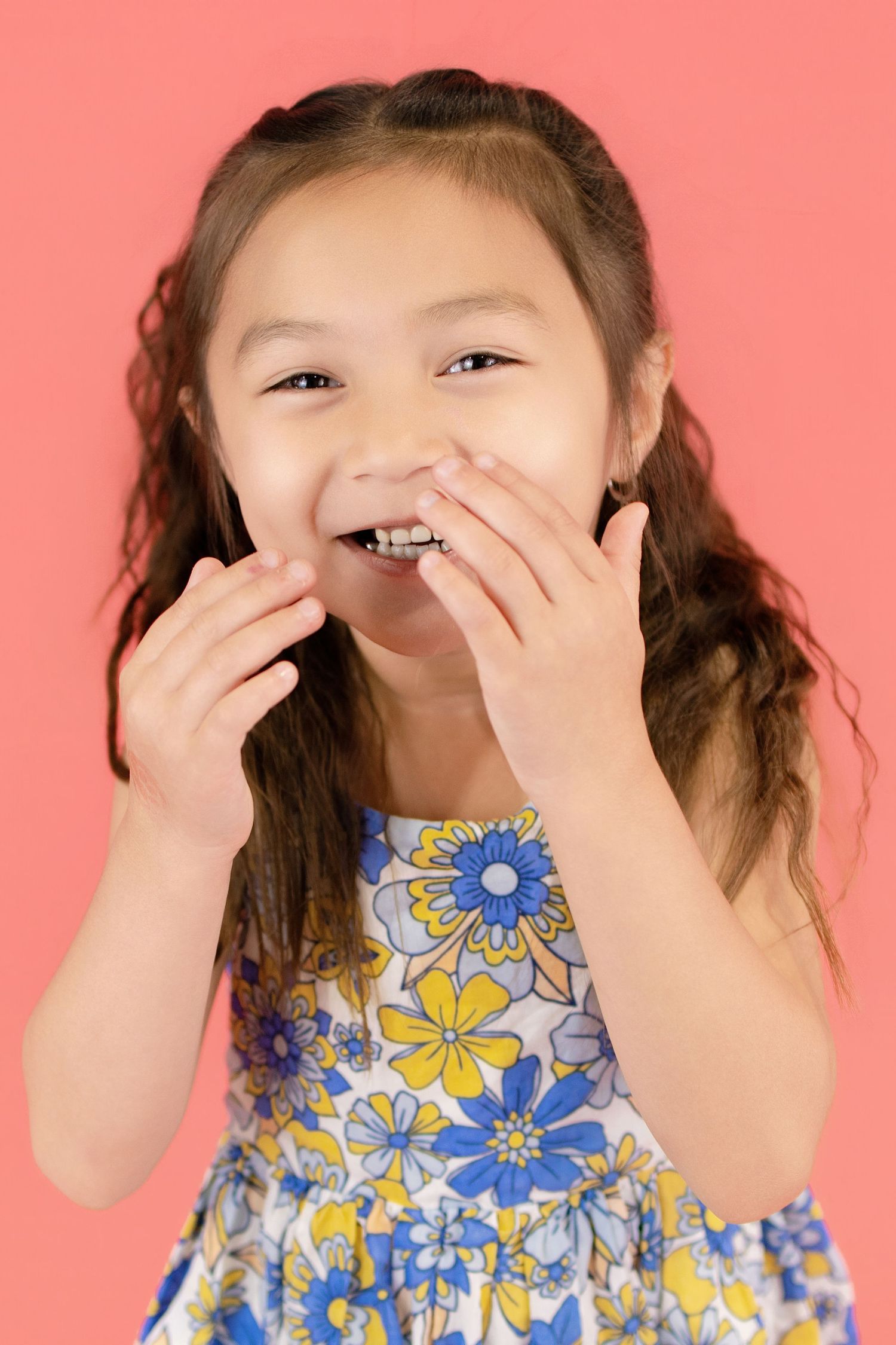 A young Asian girl in a floral dress laughs while covering her mouth with her hands against a pink background.