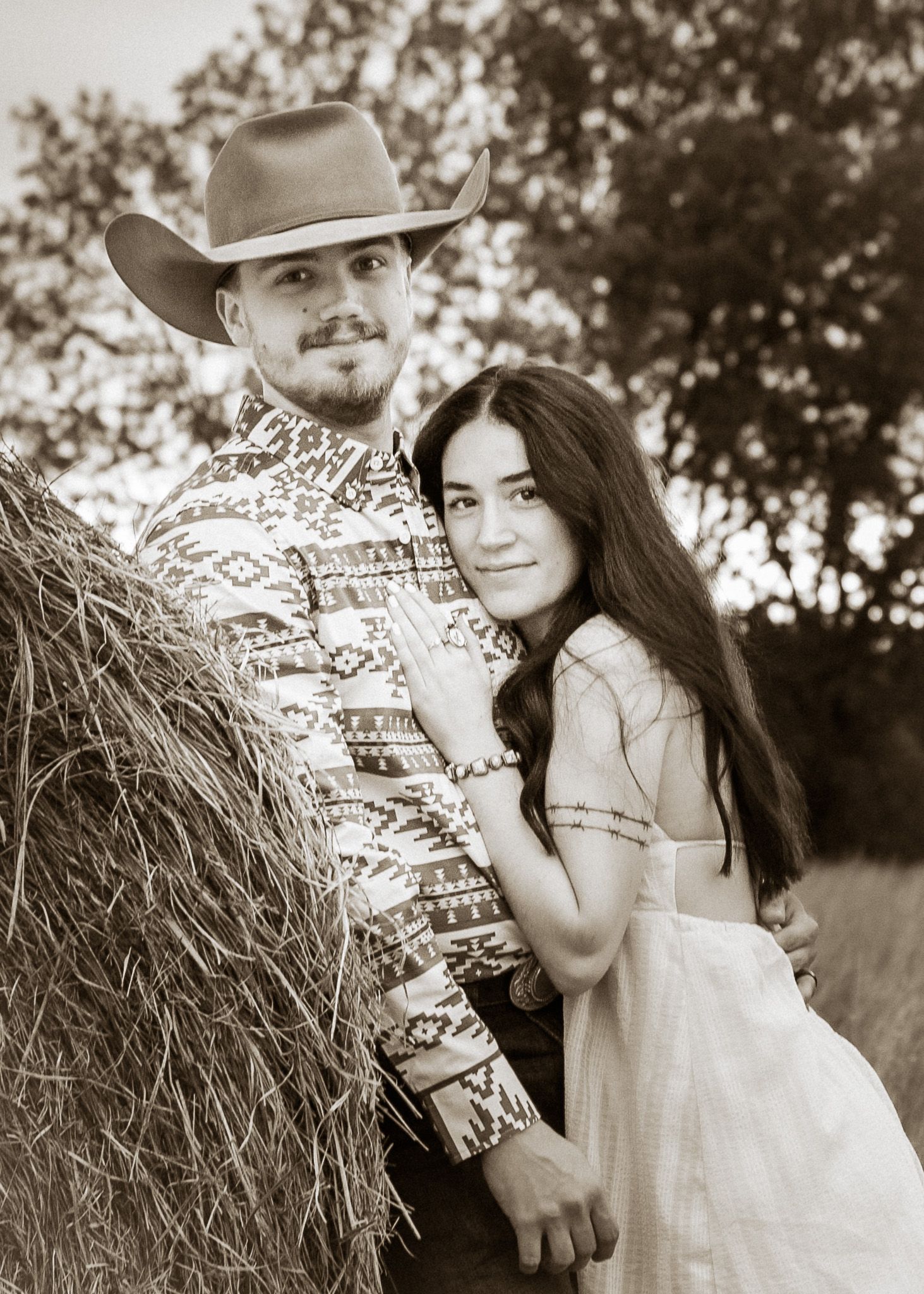 Sepia-toned photo of a couple posing by a hay bale, man in cowboy hat and patterned shirt, woman in white dress.