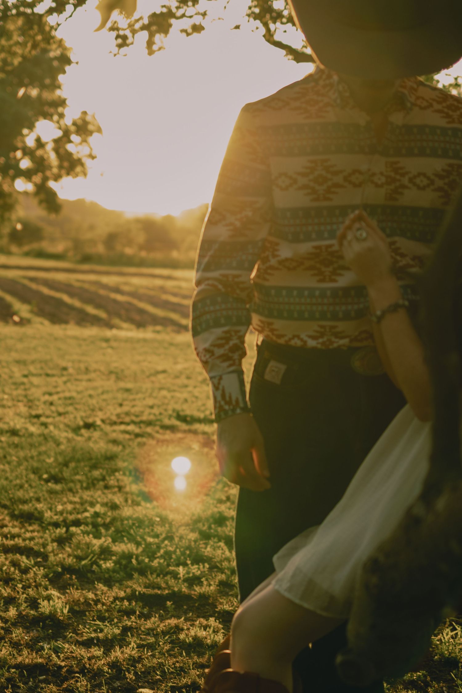 Person in patterned sweater and hat standing in sunlit field at golden hour sunset.