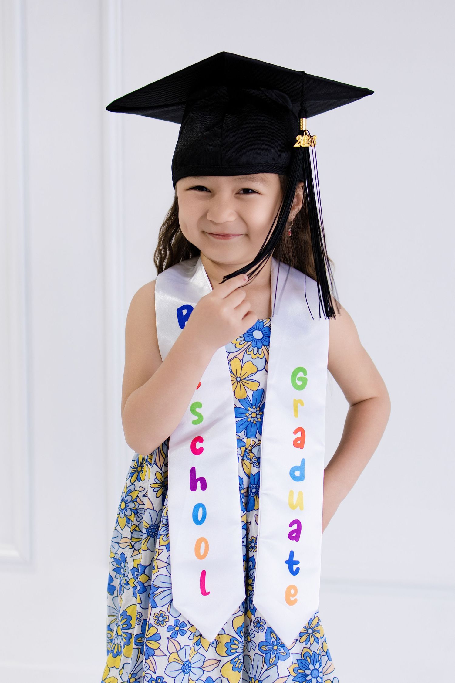 Young Asian girl wearing a preschool graduation cap and sash over a floral dress, smiling proudly.