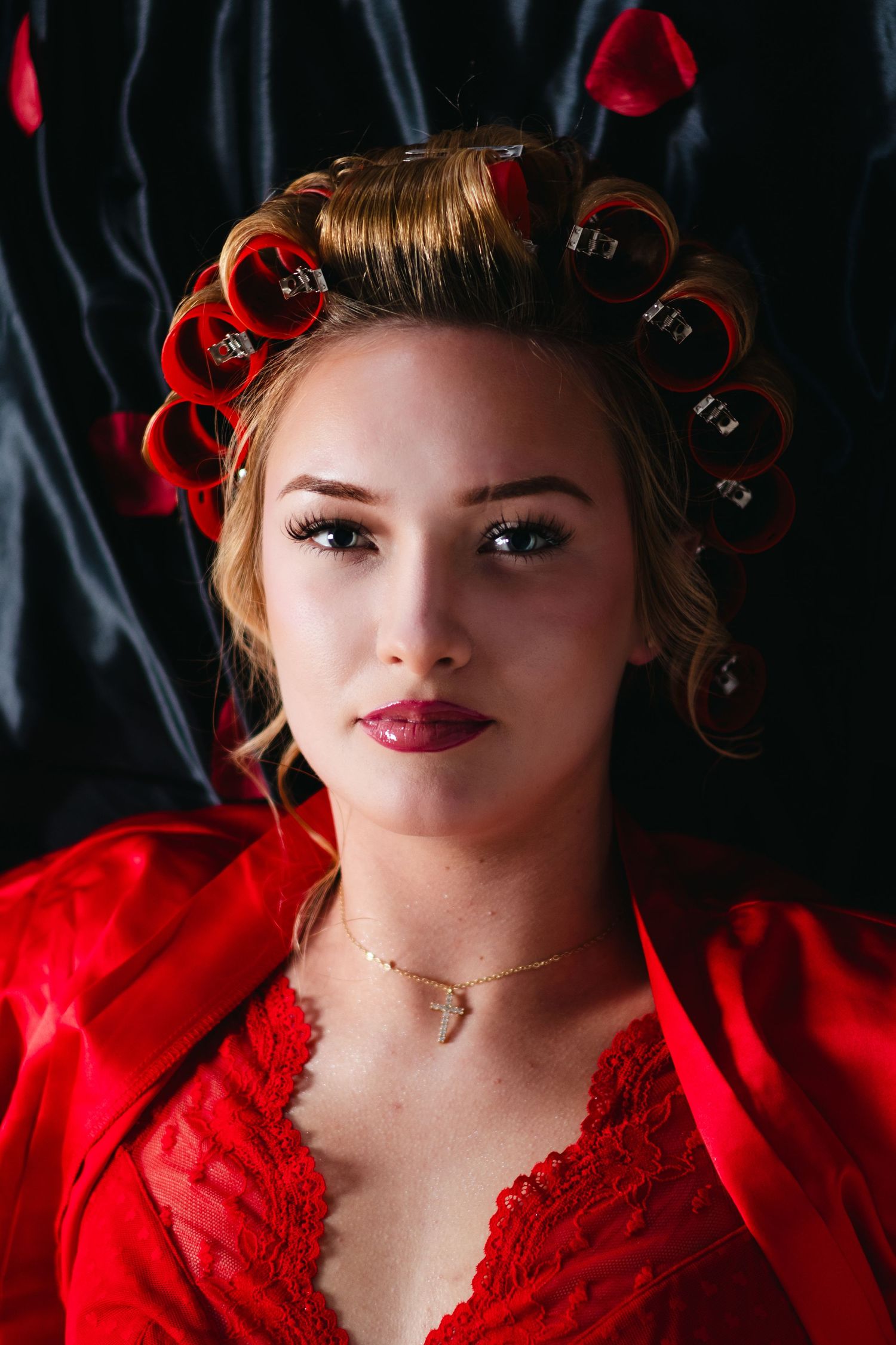 Woman with red hair curlers and red lace top lying against dark background with dramatic makeup.