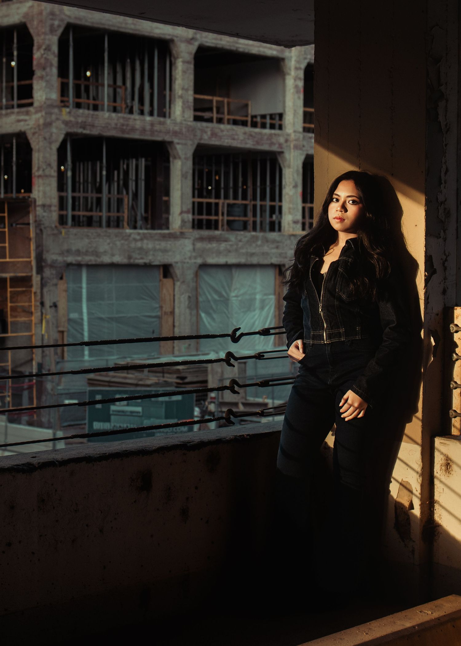 Young woman in black outfit poses on urban balcony at night with industrial building backdrop.