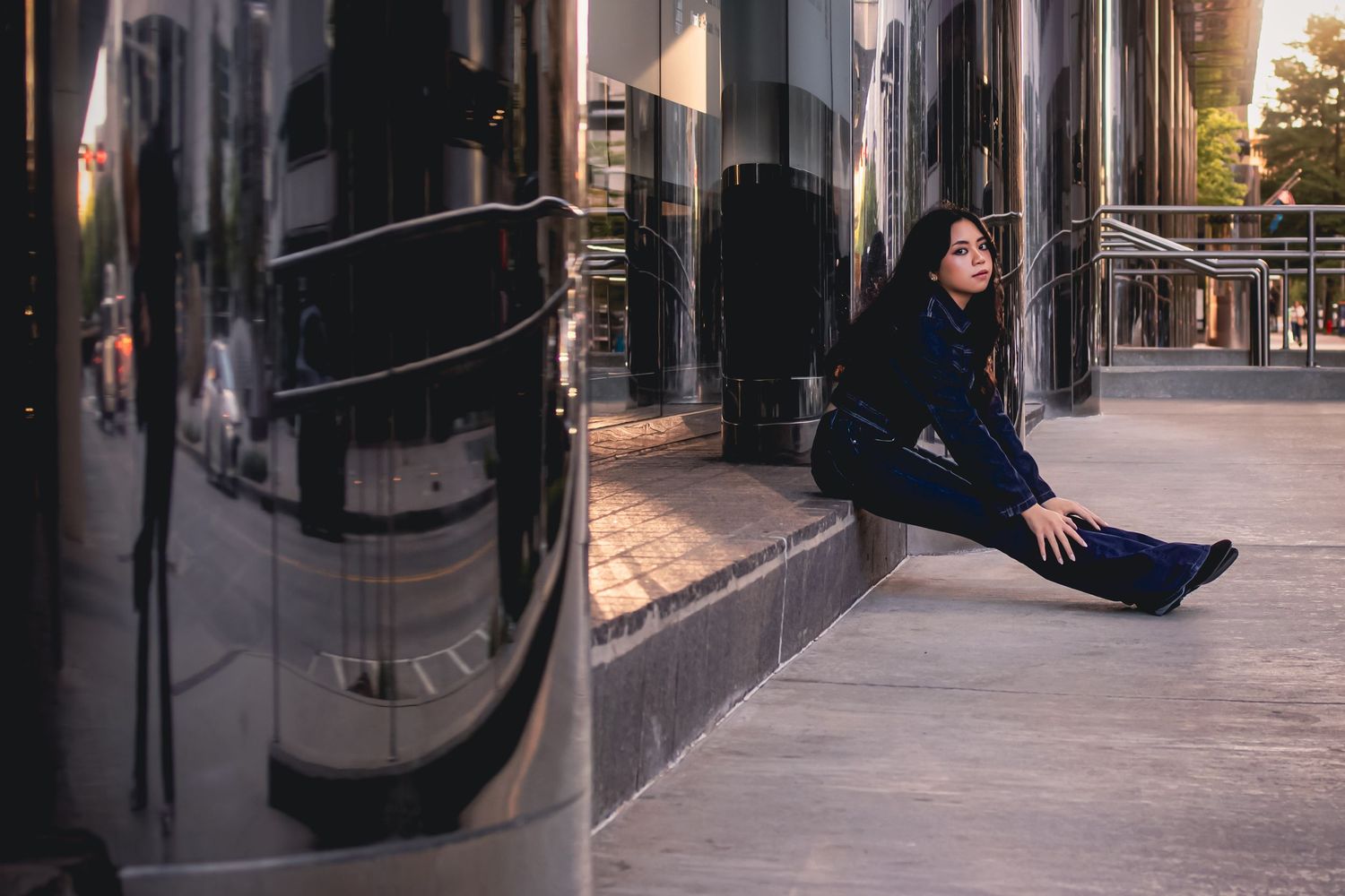 A woman in a blue outfit sits gracefully on urban steps beside reflective dark glass building columns.