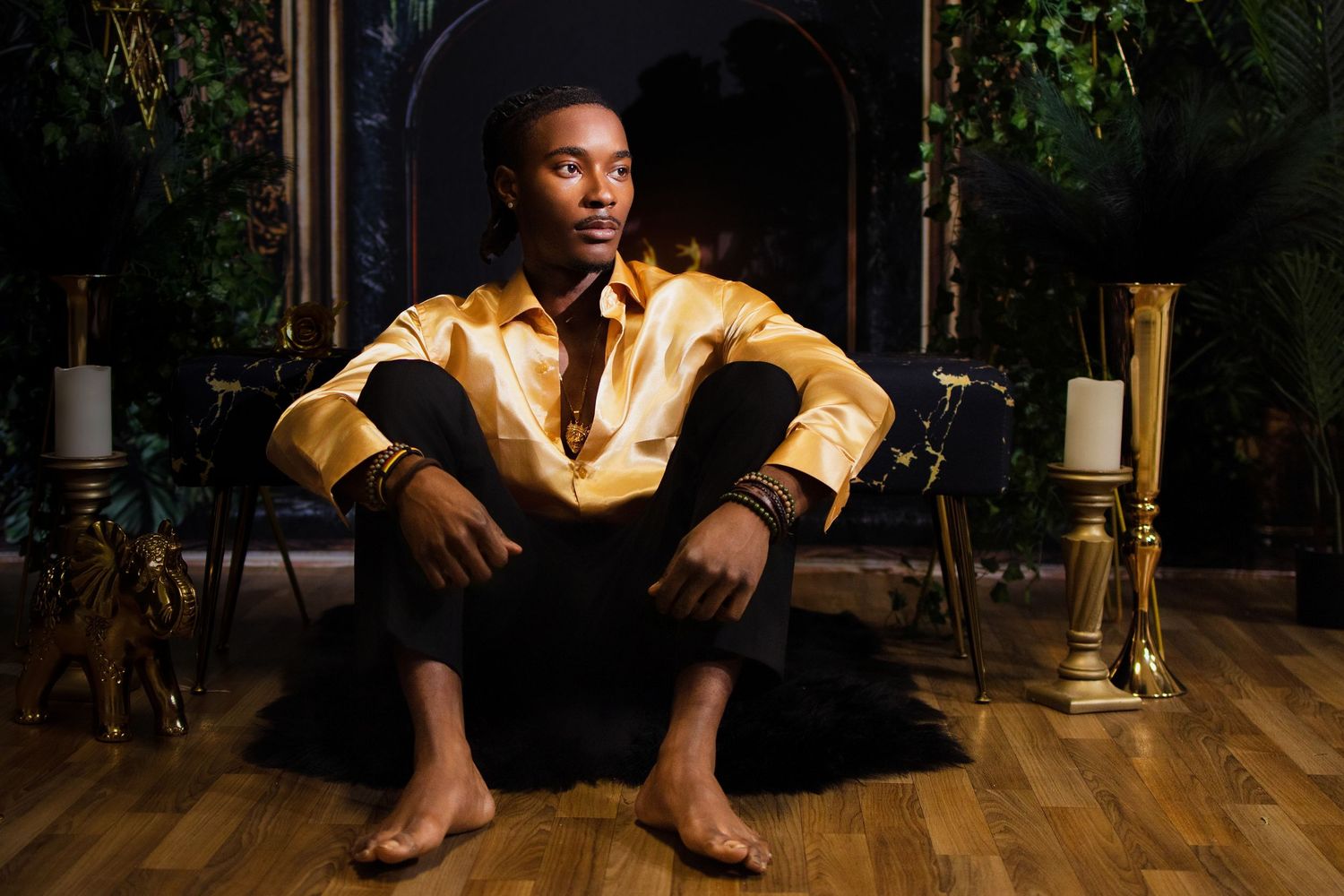 Young Black man in gold satin shirt sits barefoot on floor in dramatic candlelit dark setting.