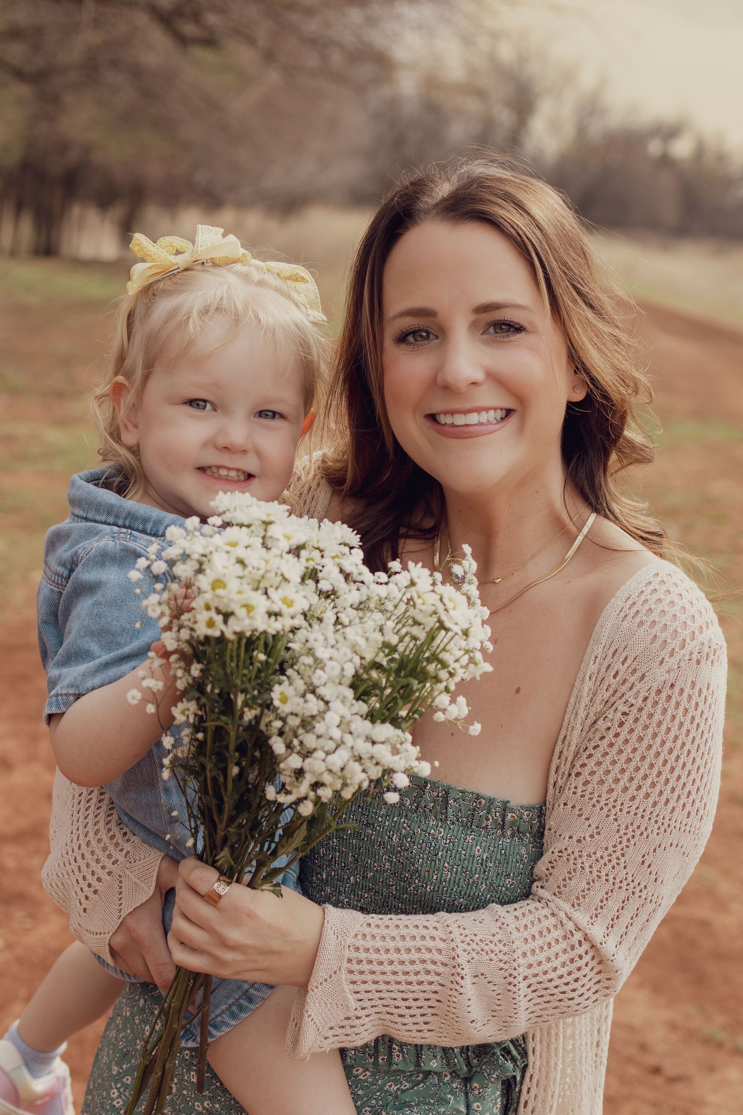 A smiling mother holds her toddler daughter while both clutch a bouquet of white wildflowers outdoors.