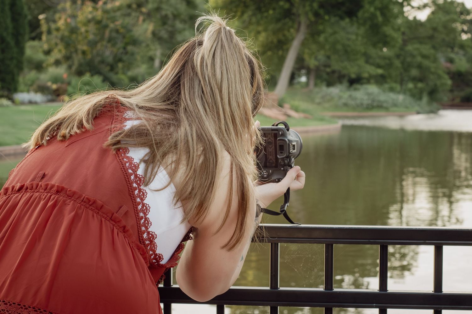 Blonde woman in red dress photographing a park pond with a vintage camera while leaning on a black railing.