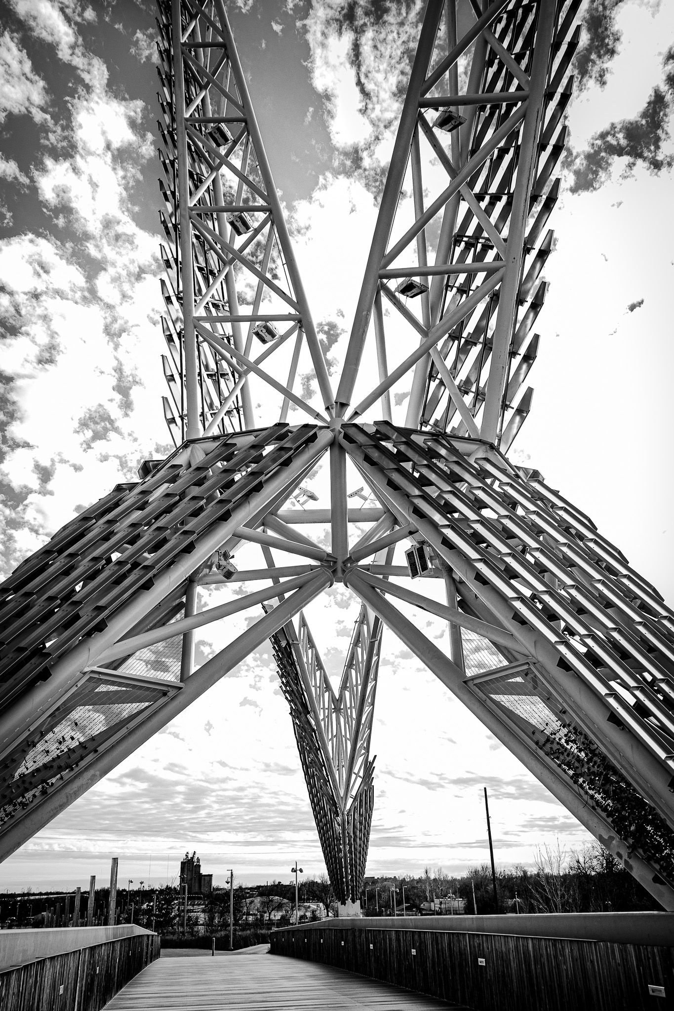 Black and white photo looking up at a massive steel truss structure against a cloudy sky with a wooden walkway below.