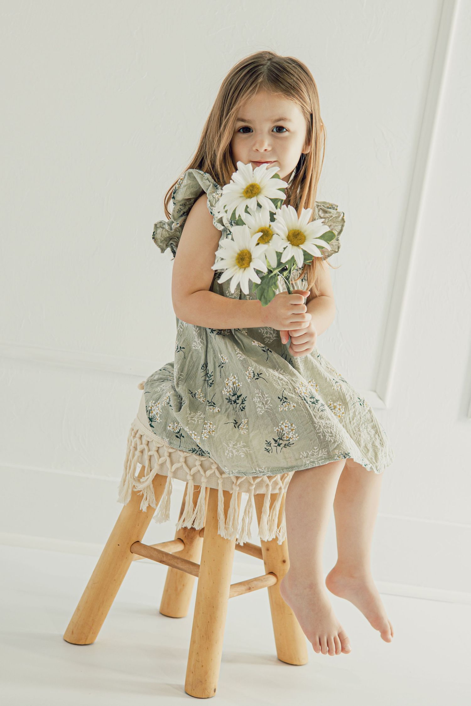 Young girl in floral green dress with fringe trim sits on wooden stool holding white daisy bouquet in white studio.