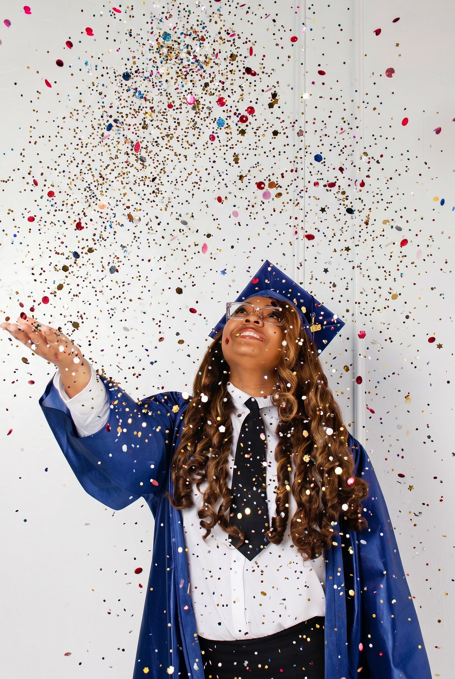 A smiling graduate in blue cap and gown tosses colorful confetti in the air, celebrating her achievement.