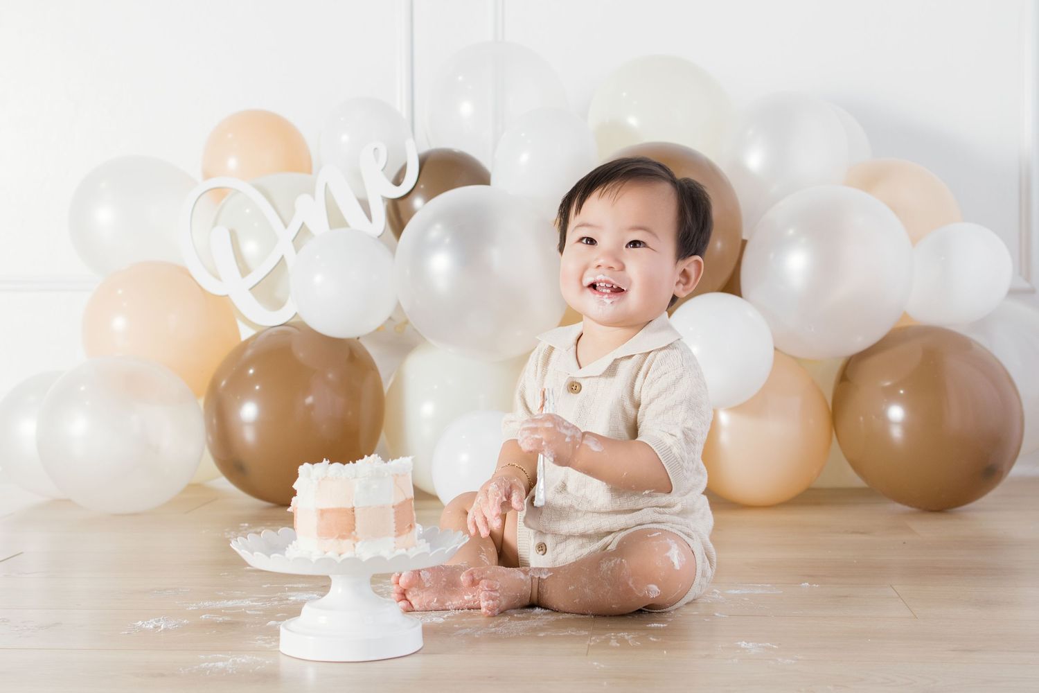 Baby smiling at first birthday cake smash with gold white balloons and One sign backdrop.