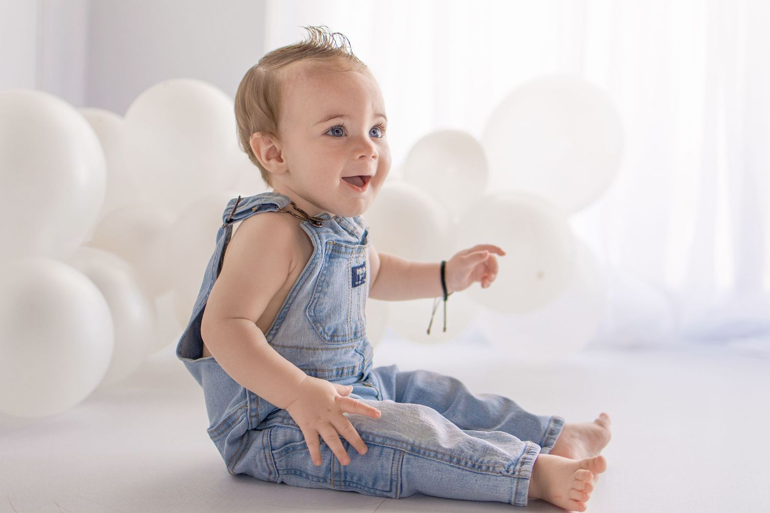 Smiling baby in denim overalls sitting on white floor surrounded by white balloons in a bright studio setting.