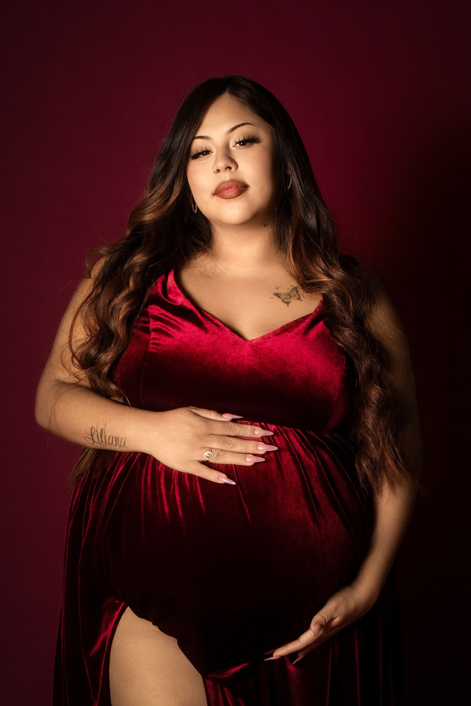 Pregnant woman in red velvet gown cradling baby bump against dark red background in elegant maternity portrait.