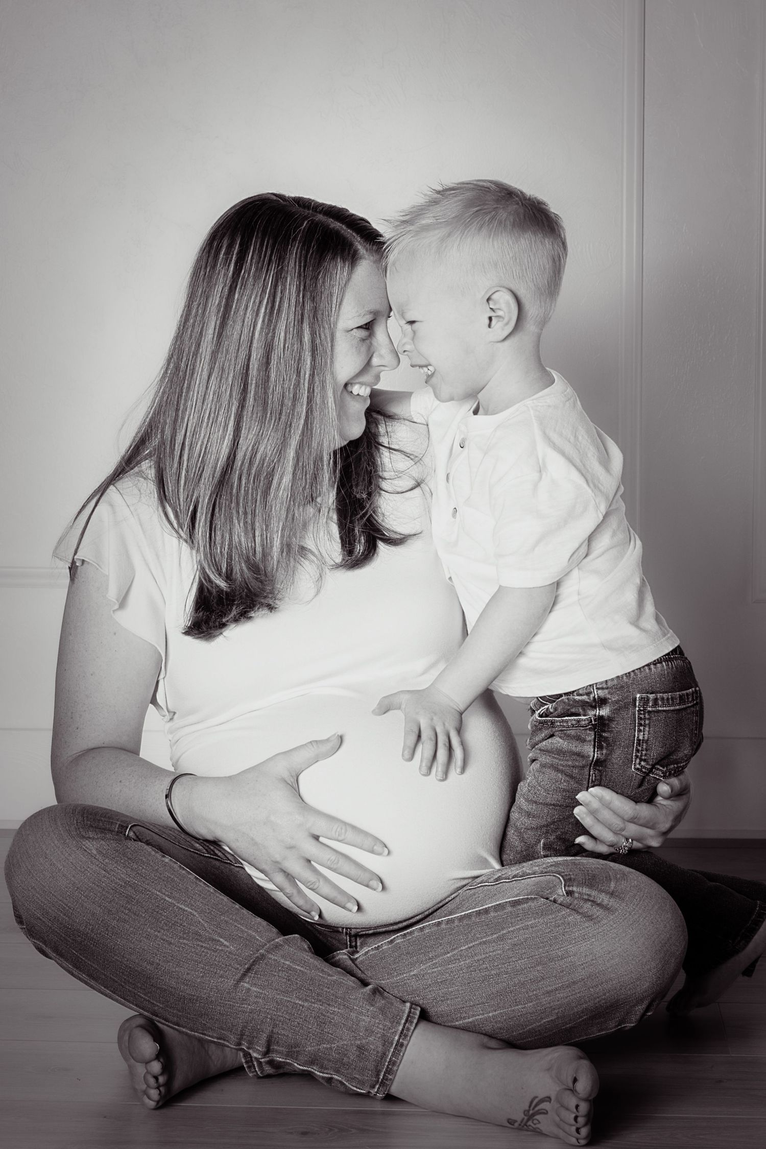Black and white photo of a pregnant mother sitting cross-legged on floor, smiling with her young blonde son.