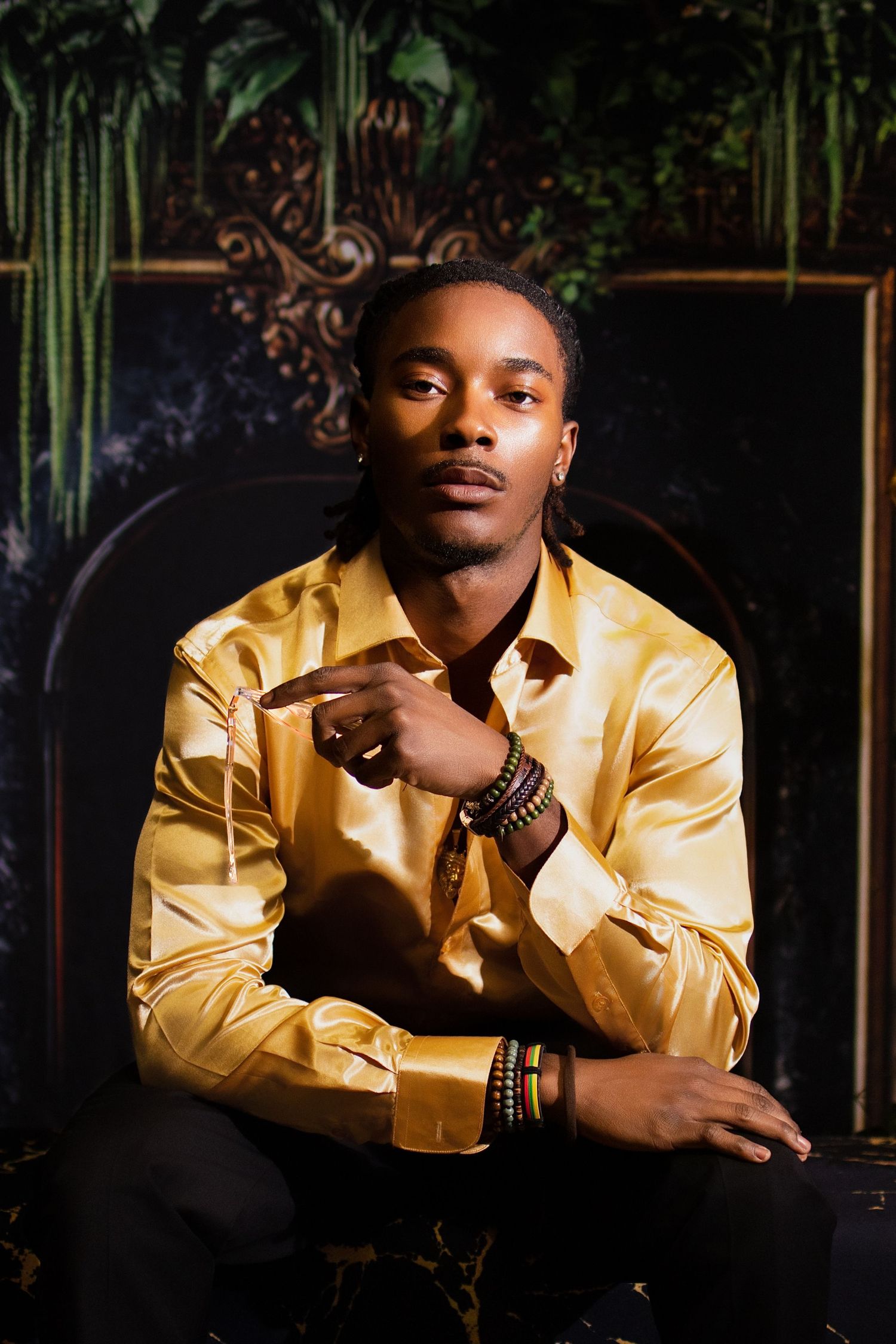 Stylish man in gold satin shirt with bracelets sits confidently pointing finger in dramatic dark setting.