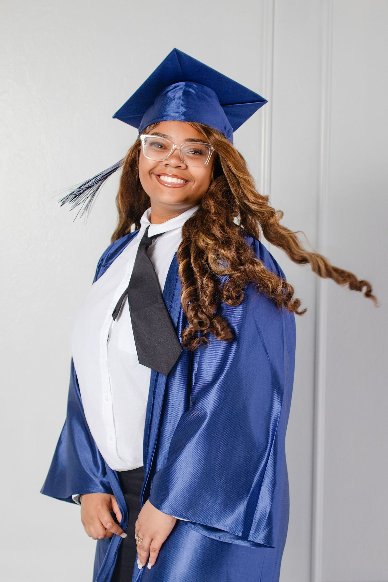 Smiling young woman in blue graduation cap and gown with glasses and curly hair posing for graduation photo.