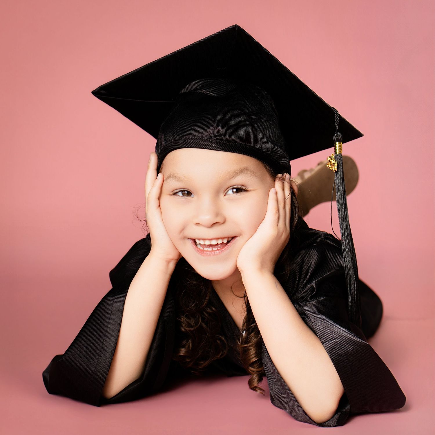 Young girl in black graduation cap and gown smiling with hands on cheeks against pink background.