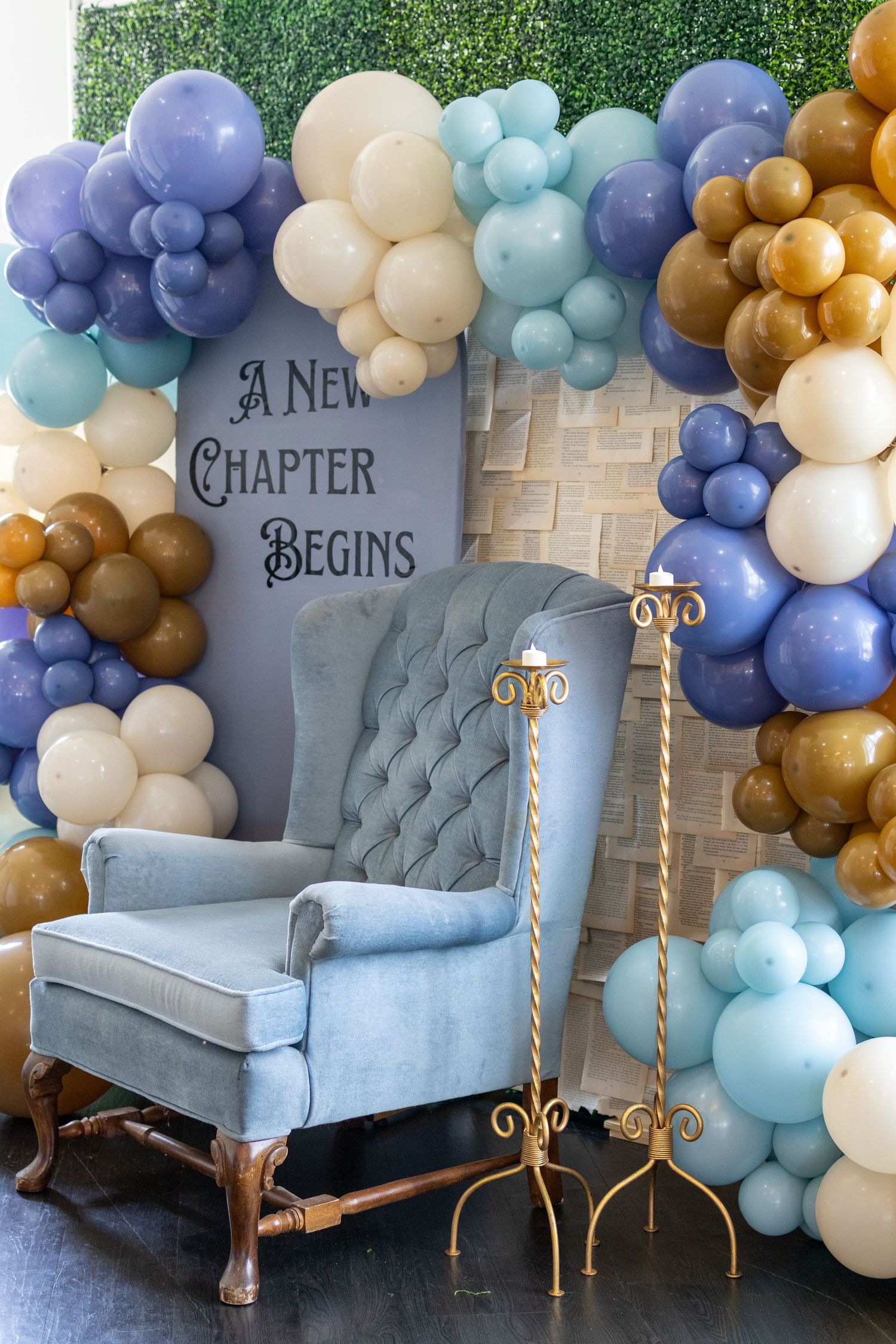 Elegant blue velvet chair surrounded by balloon garland in blue, gold, and cream with 'A New Chapter Begins' sign.