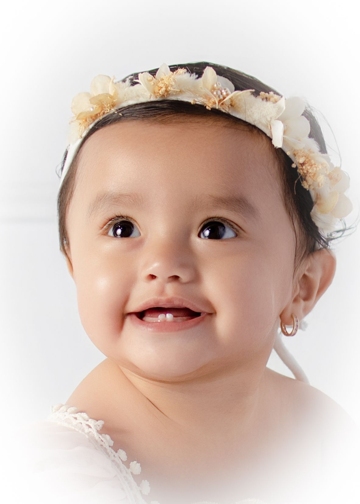 Adorable smiling baby girl wearing a floral crown headband and small hoop earrings, with bright brown eyes.