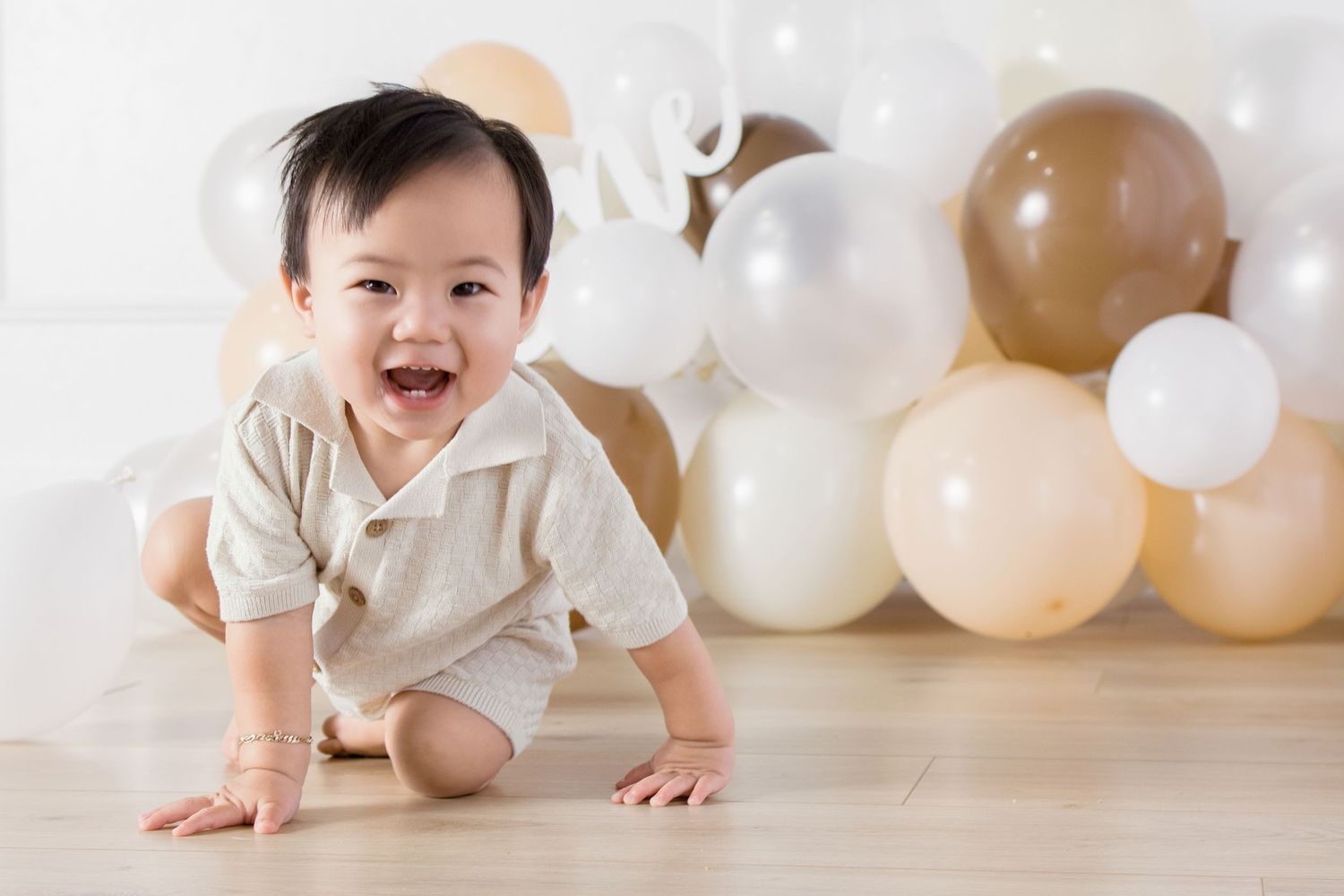Smiling baby crawling on hardwood floor with neutral balloons and white decor in background.
