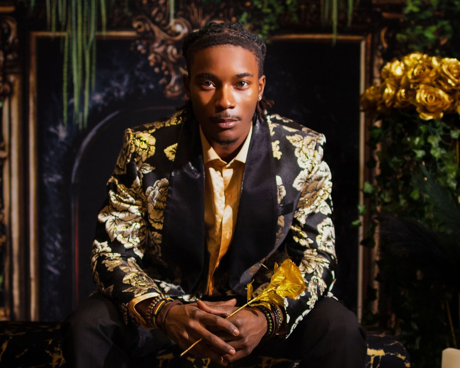 Young man in gold floral blazer poses confidently in elegant dark setting with lush greenery.