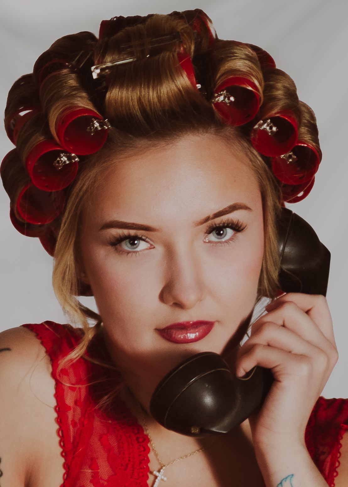 Woman with red hair rollers holding a vintage black telephone, wearing red lace, retro pin-up style portrait.