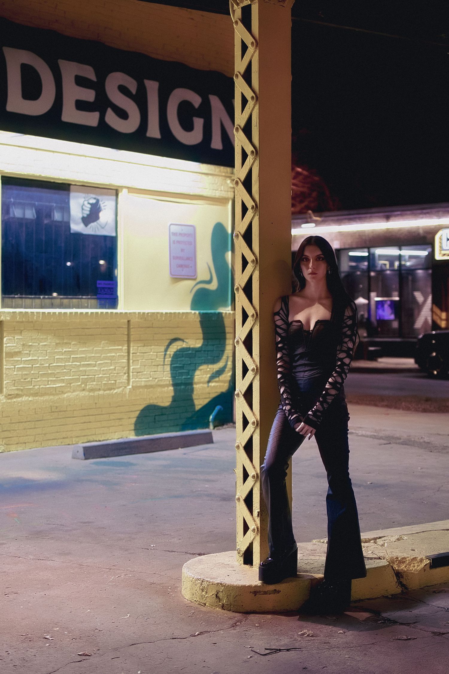 Woman in black corset and flare pants posing at night under Design sign.