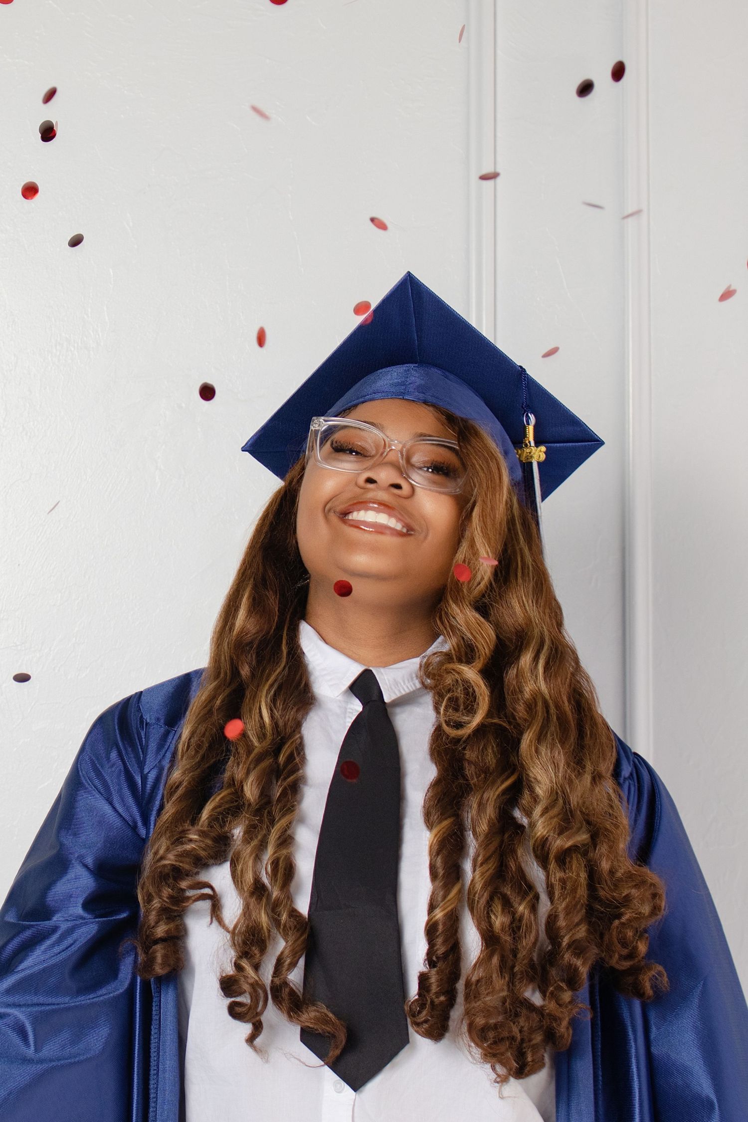 Smiling graduate in blue cap and gown with glasses and black tie, celebrating with falling confetti.