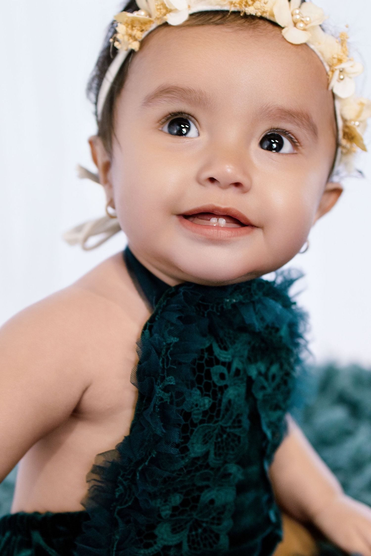Adorable baby girl in a dark green lace dress and floral headband smiling sweetly at the camera.