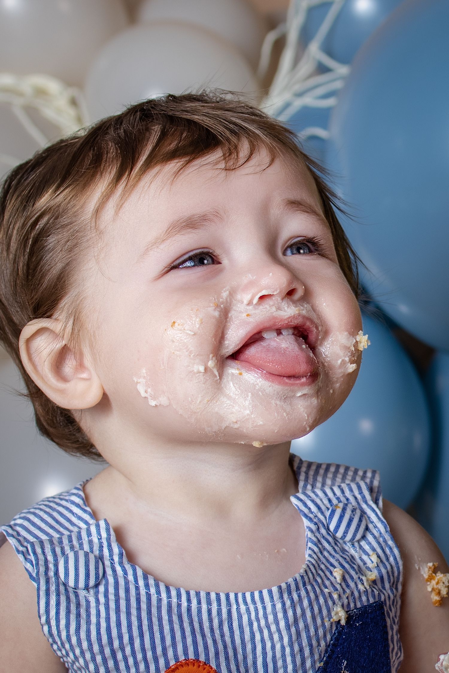 Adorable baby with frosting-covered face smiles with tongue out during first birthday cake smash with blue balloons.