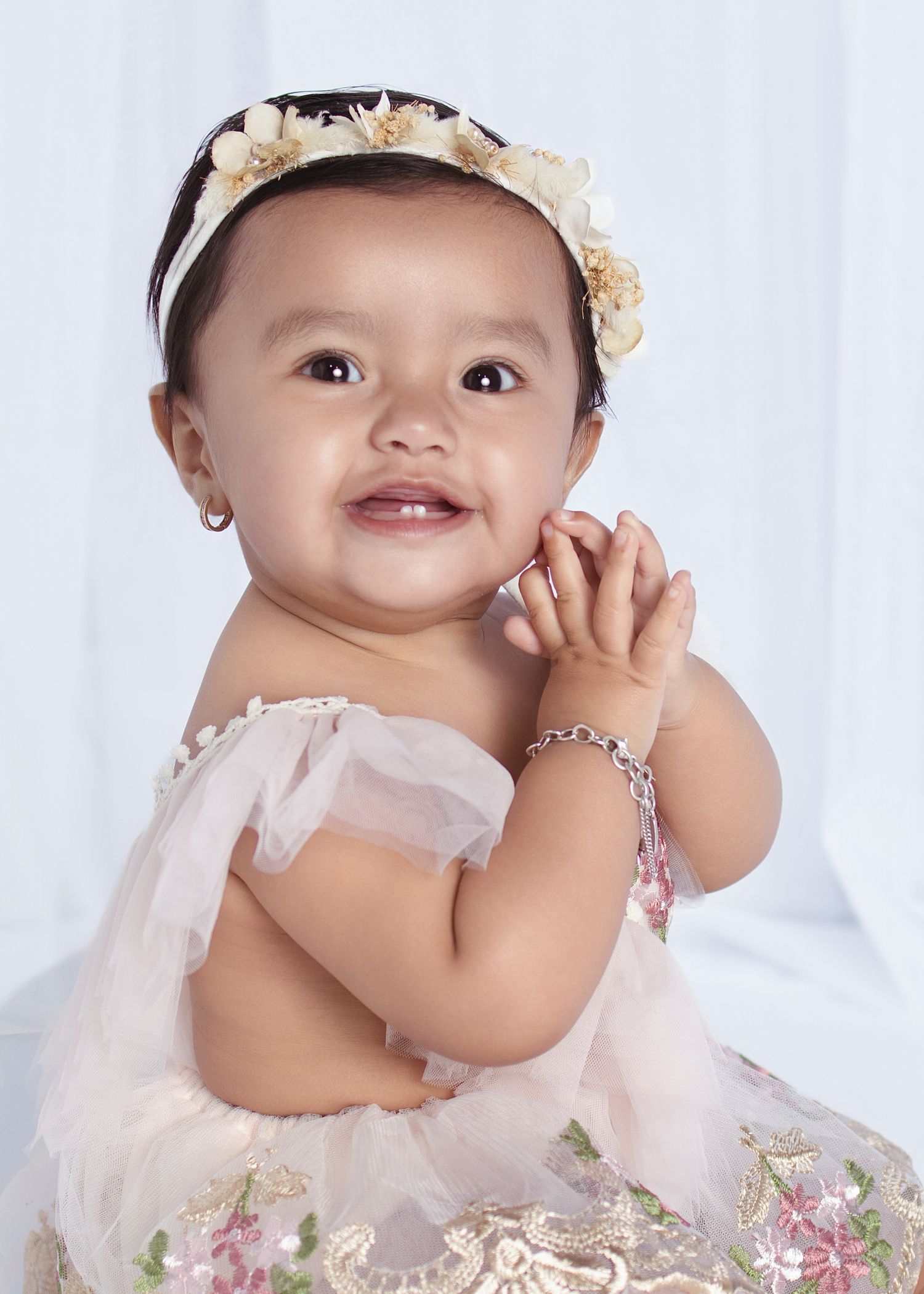 Smiling baby girl in floral dress and headband, wearing pearl earrings and bracelet, clapping hands.