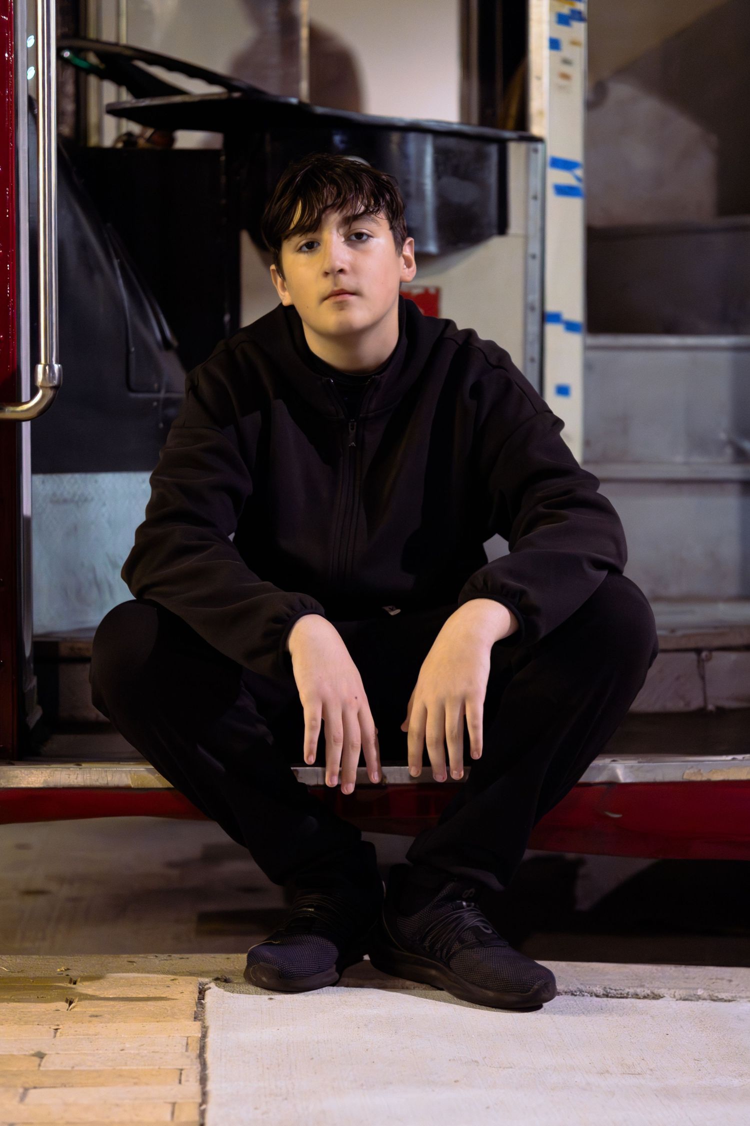 Young teen in all-black hoodie and sneakers sitting outdoors at night with a confident pose.