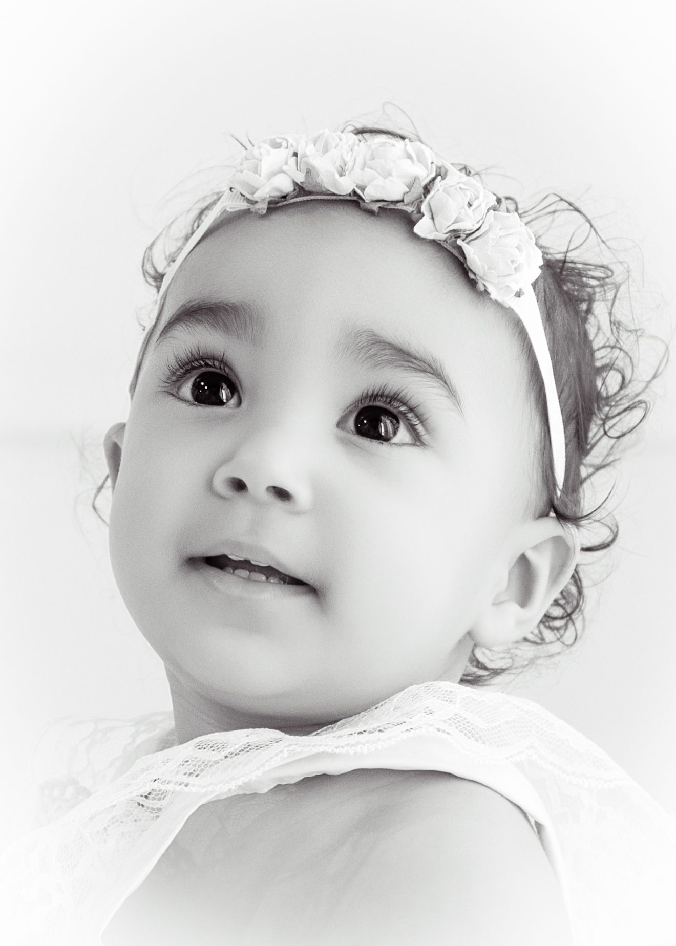Black and white portrait of a cute baby girl wearing a floral headband, looking up with wide curious eyes.