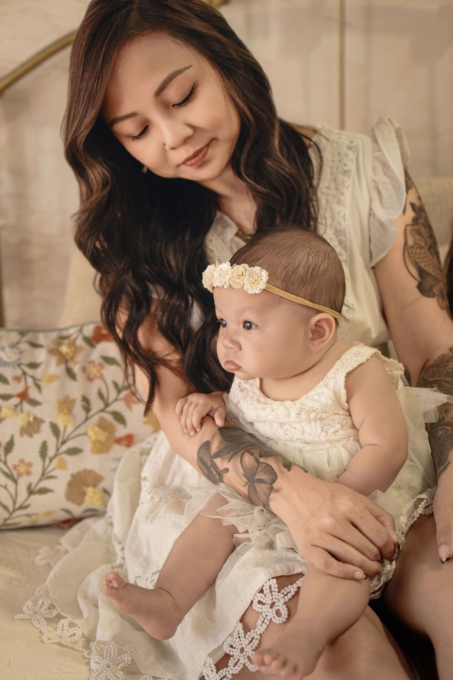 A young Asian mother with tattoos tenderly holds her baby girl in a floral headband, both dressed in white lace.