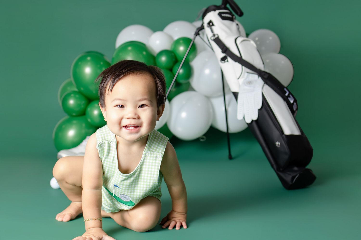 Smiling baby in green romper poses with golf bag and green and white balloon garland on green background.