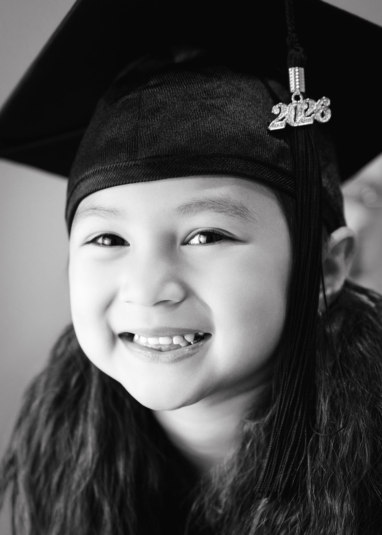 Smiling young girl wearing a black graduation cap with a 2023 tassel charm, black and white portrait photo.