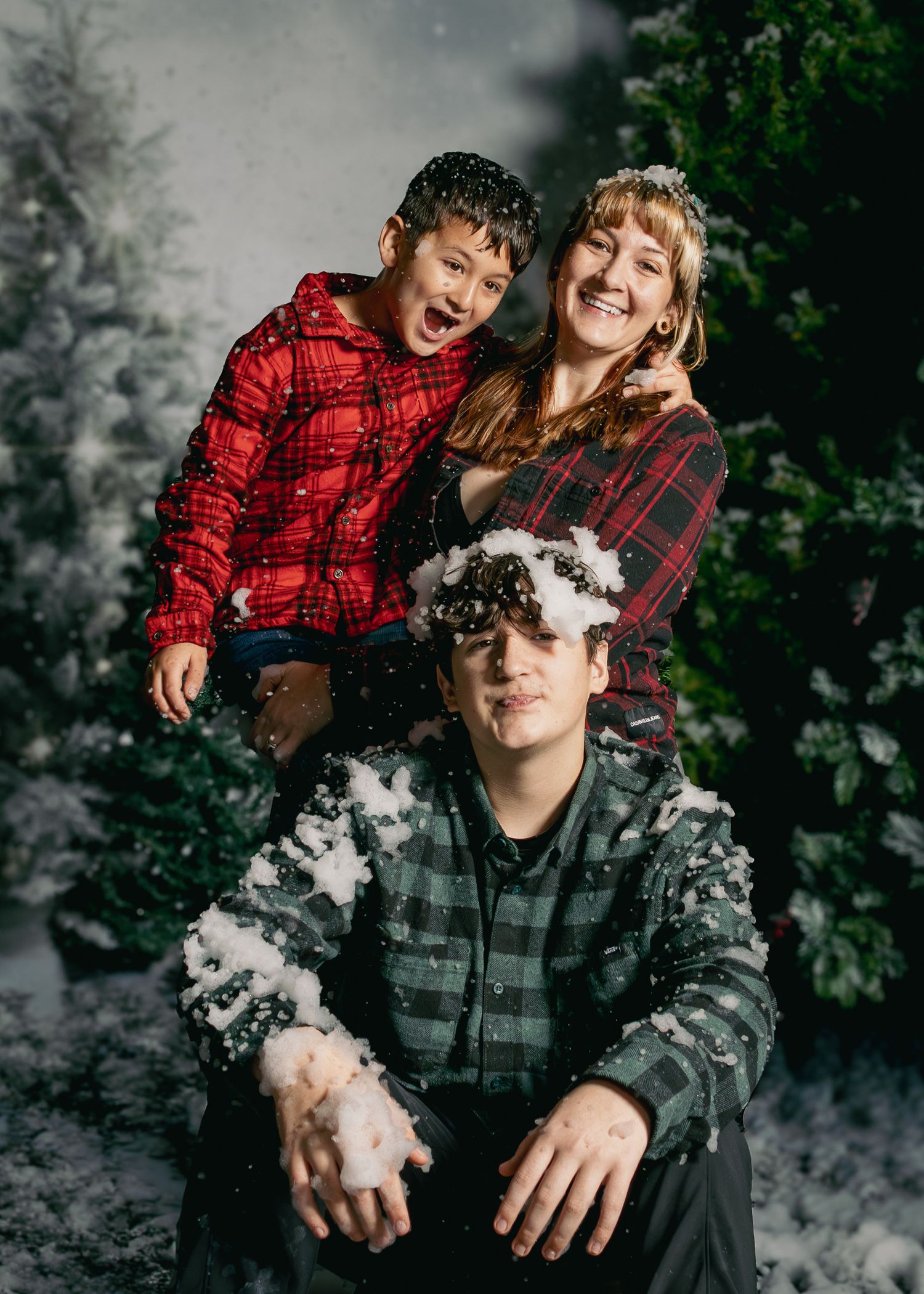 Family of three posing in a snowy Christmas photo studio with festive plaid outfits and snow-covered evergreen trees.