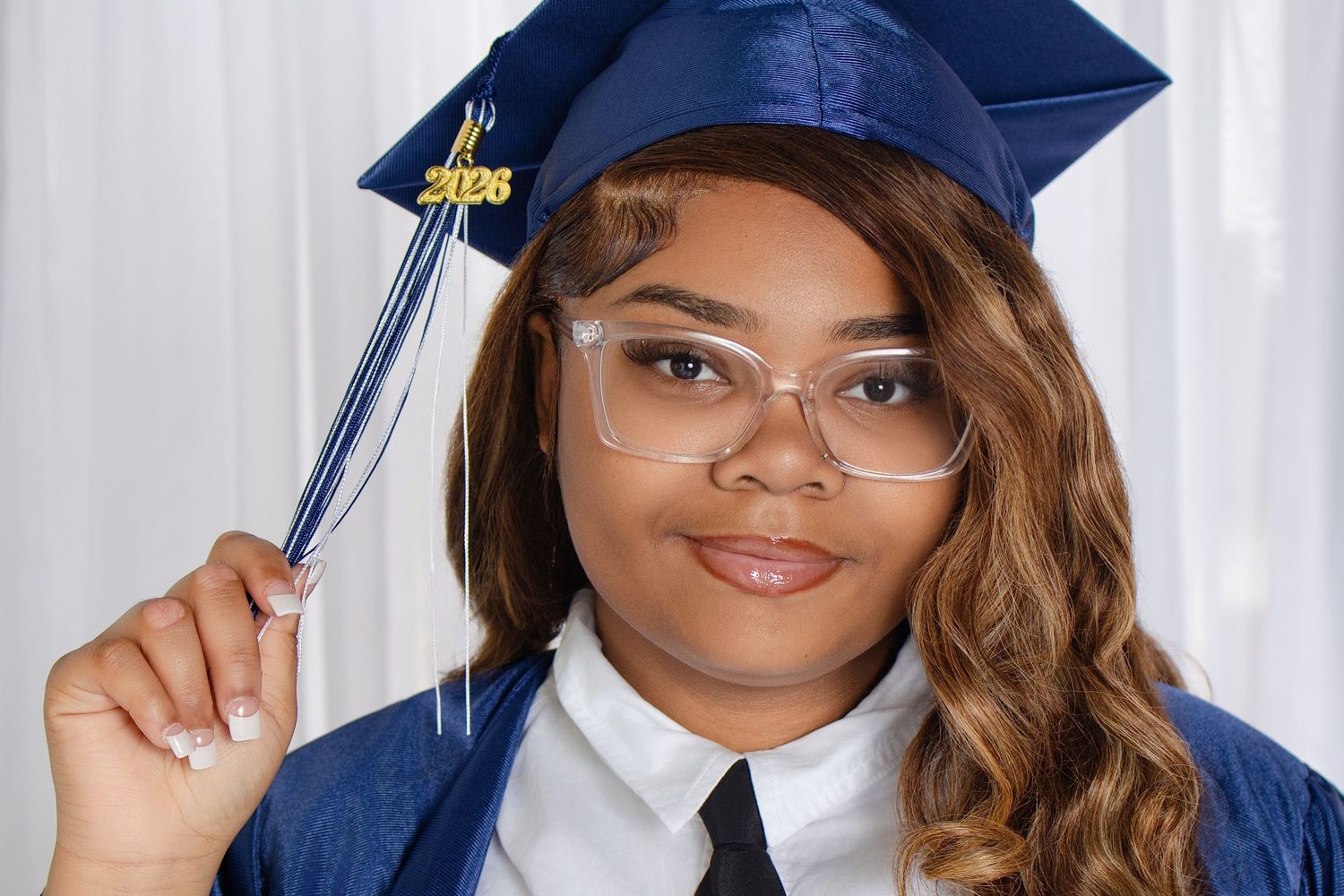 A young woman in a blue graduation cap and gown holds her tassel while wearing clear-framed glasses.