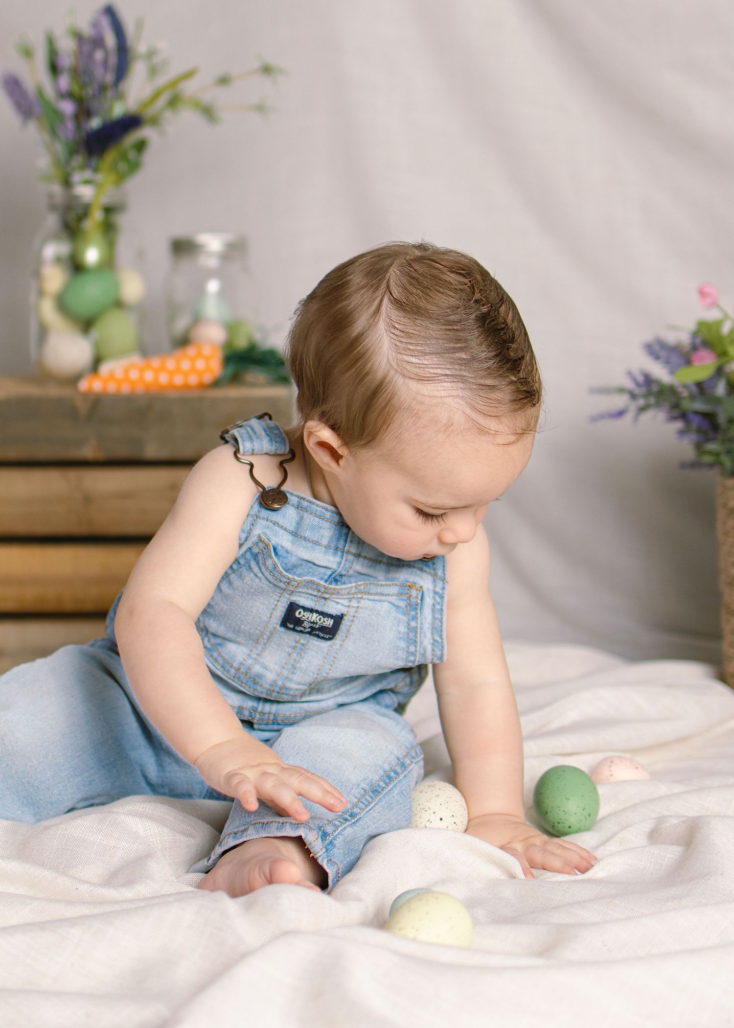 Curious baby in denim overalls playing with colorful Easter eggs on white fabric backdrop.