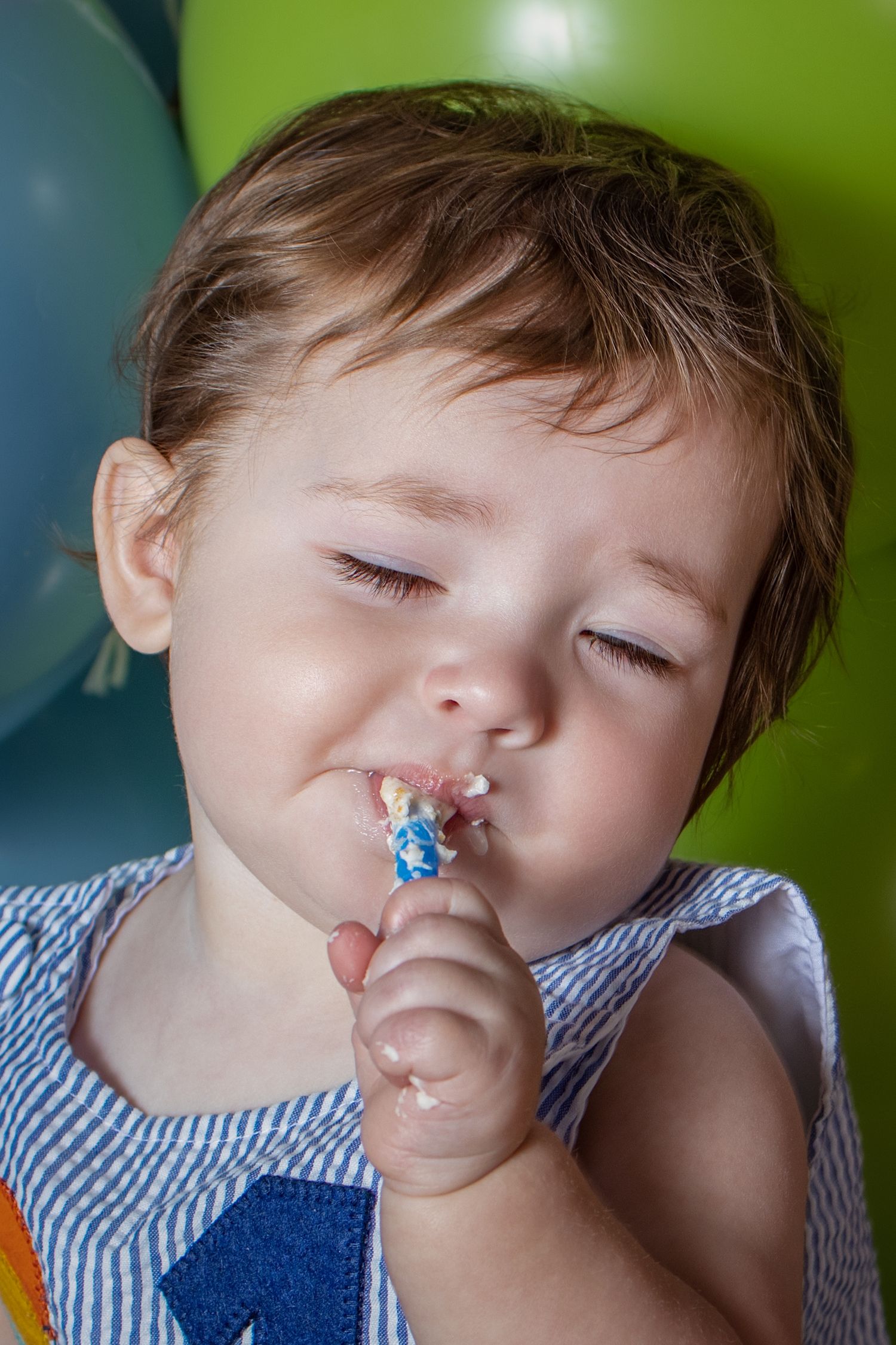Messy baby with frosting on face licking a birthday cake topper at a first birthday party with colorful balloons.