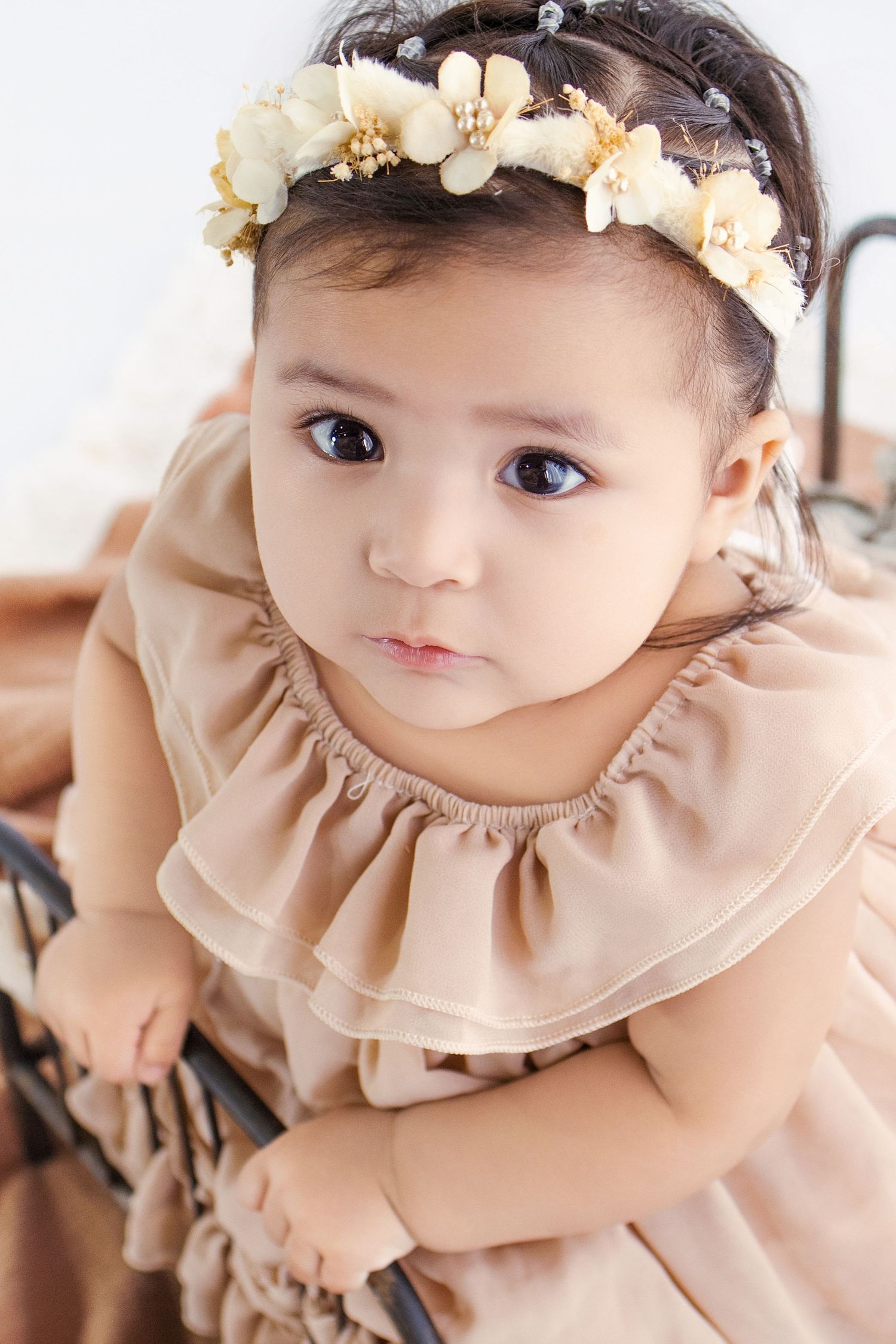Adorable baby girl in beige ruffled dress and floral crown gazing up with big dark eyes.