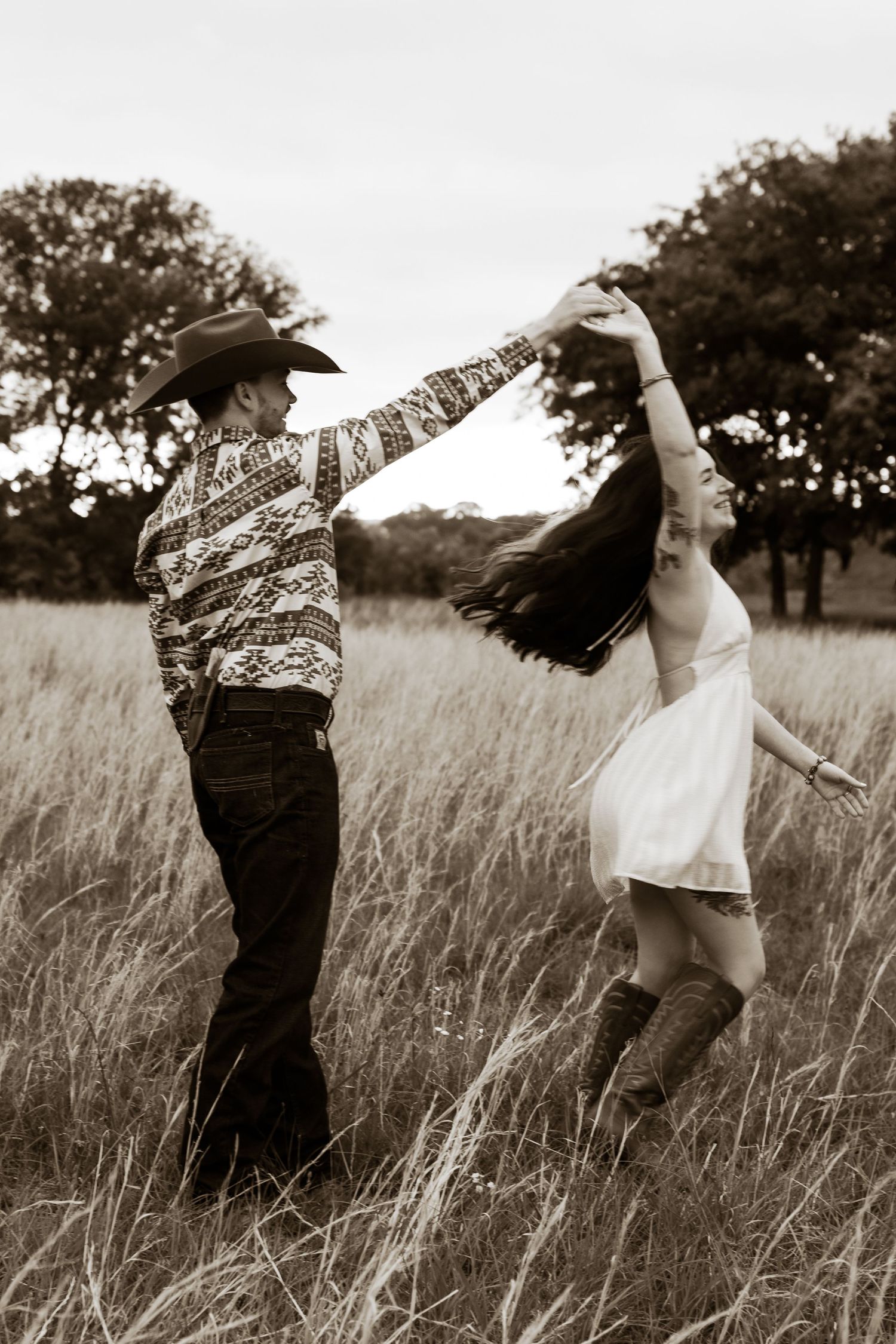 Sepia toned photo of couple dancing in field, man in cowboy hat spinning woman in white dress and boots.