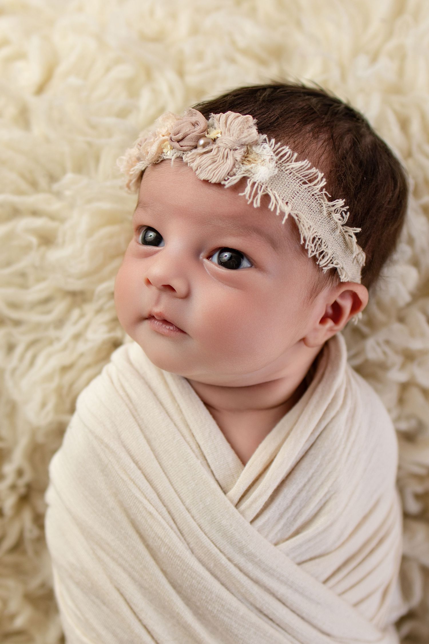 Newborn baby girl swaddled in cream wrap with floral headband, lying on fluffy white backdrop.